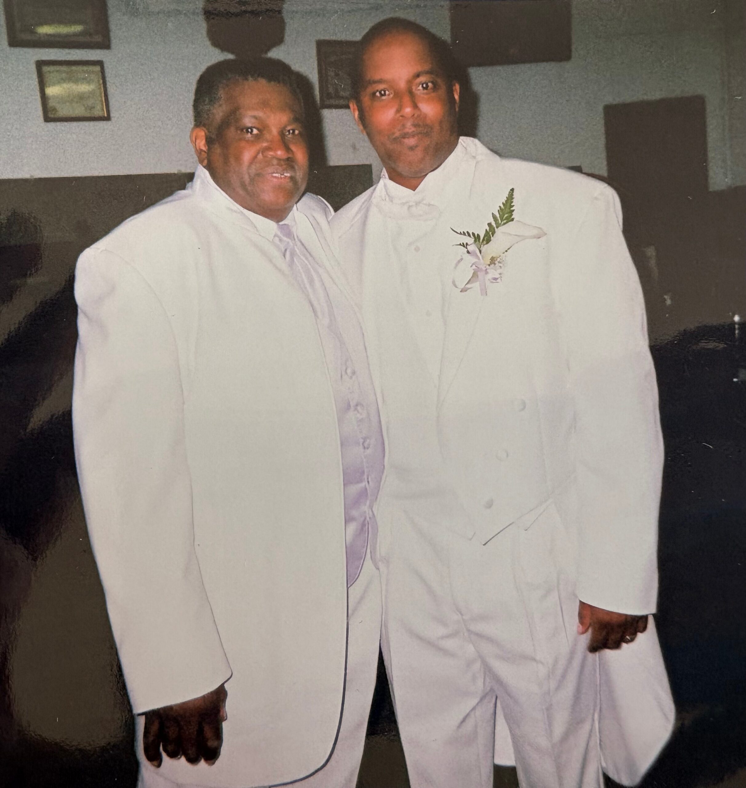 Two men in white suits posing together at a formal event, one with a boutonniere.