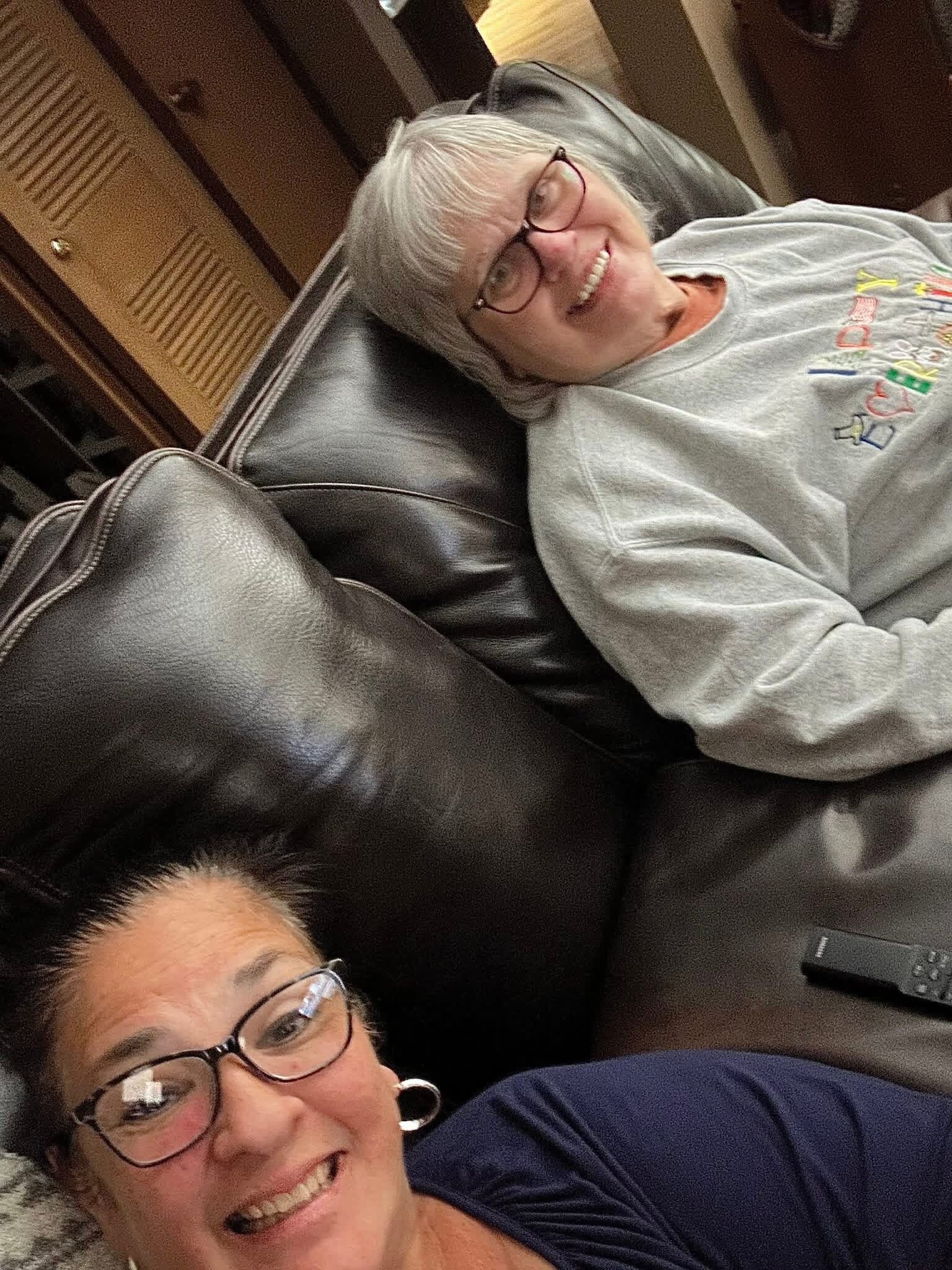 Smiling woman with short gray hair and glasses leaning on a dark leather couch, wearing a light gray sweatshirt with colorful graphics.