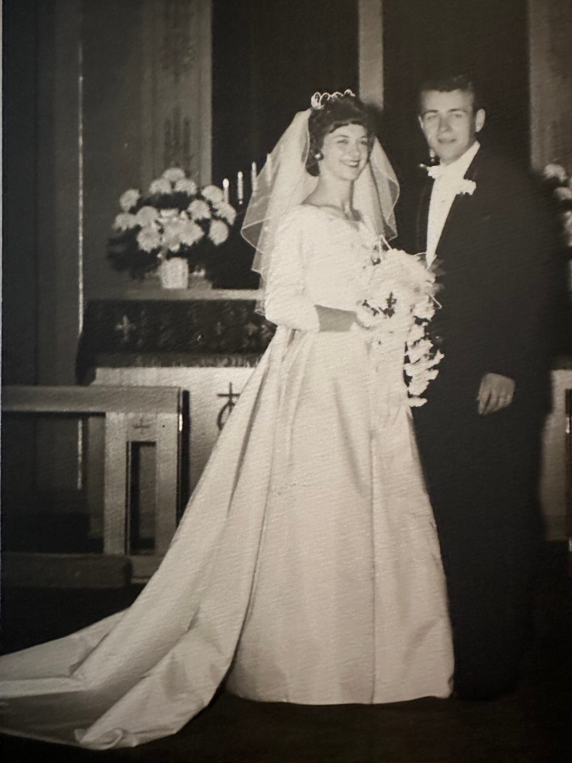 Bride in a long wedding dress and veil, holding a bouquet, stands beside a groom in a tux at a church ceremony.