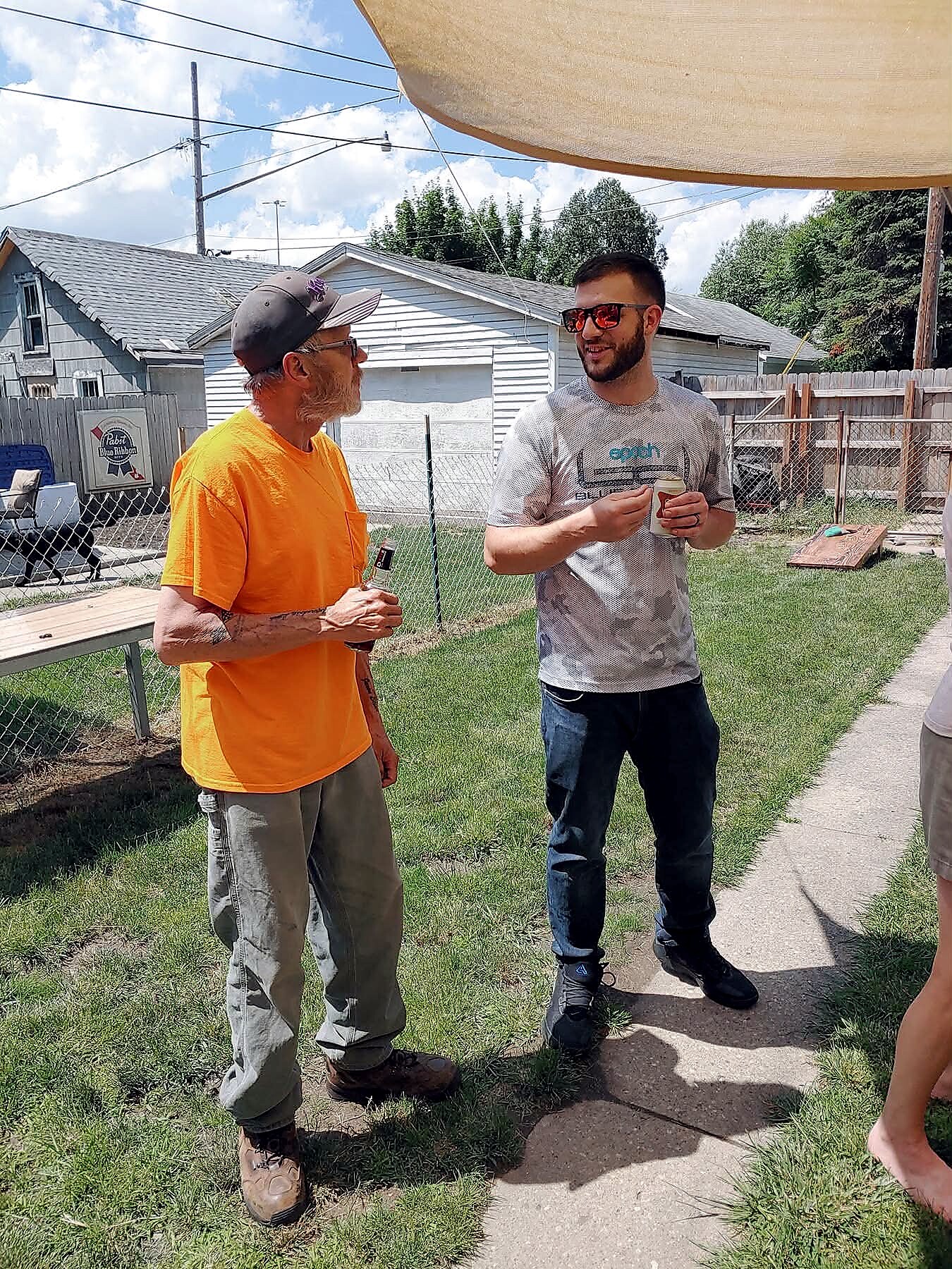 Two men stand and chat in a sunny backyard; one wears an orange shirt and cap, the other a gray T‑shirt with red sunglasses, holding a cup. Two men outdoors near a chain-link fence and white houses; one in an orange shirt and cap, the other in a gray shirt with red sunglasses. Casual conversation between two men in a backyard under a shade sail, both holding cups. Informal outdoor chat between friends in a sunny suburban yard.