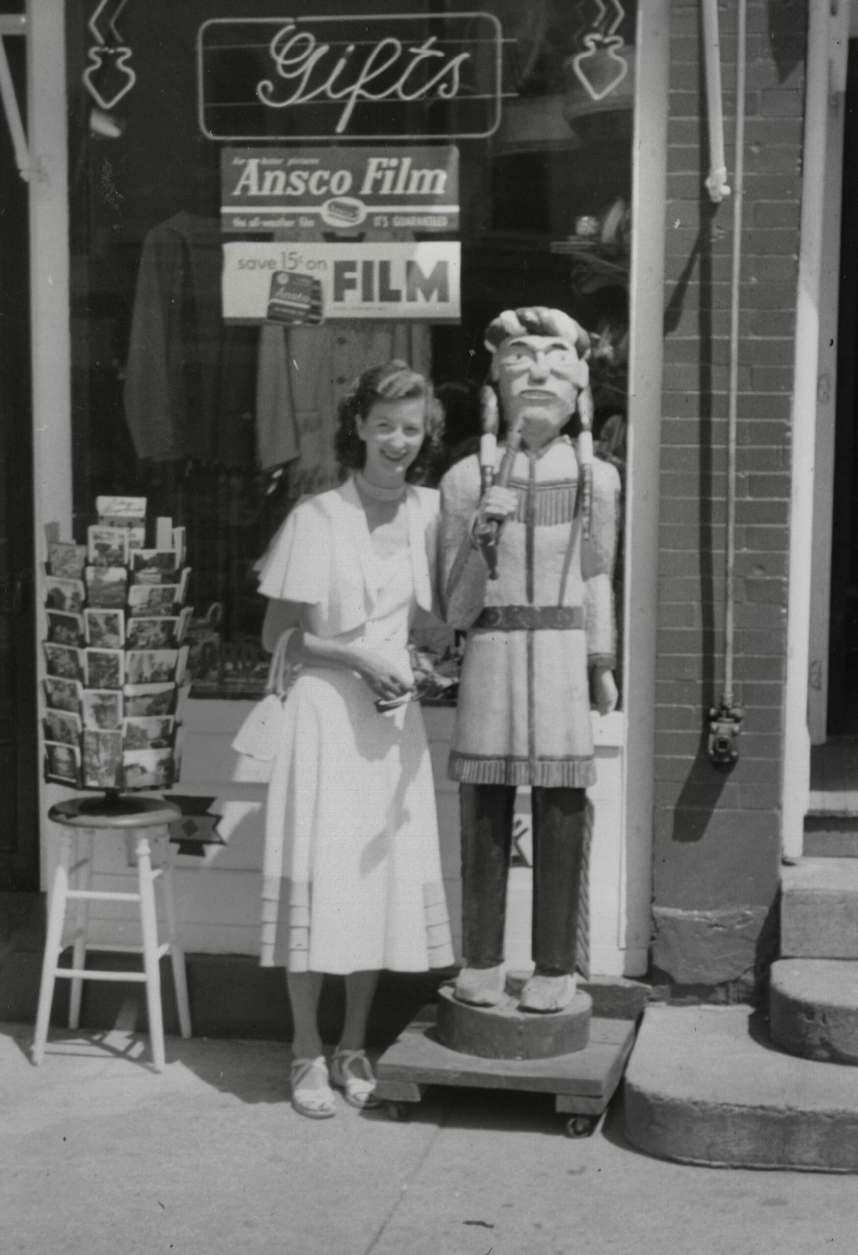 Girl in a light dress posing beside a life-sized Native American statue outside a gift shop window with a 'Gifts' sign above them.