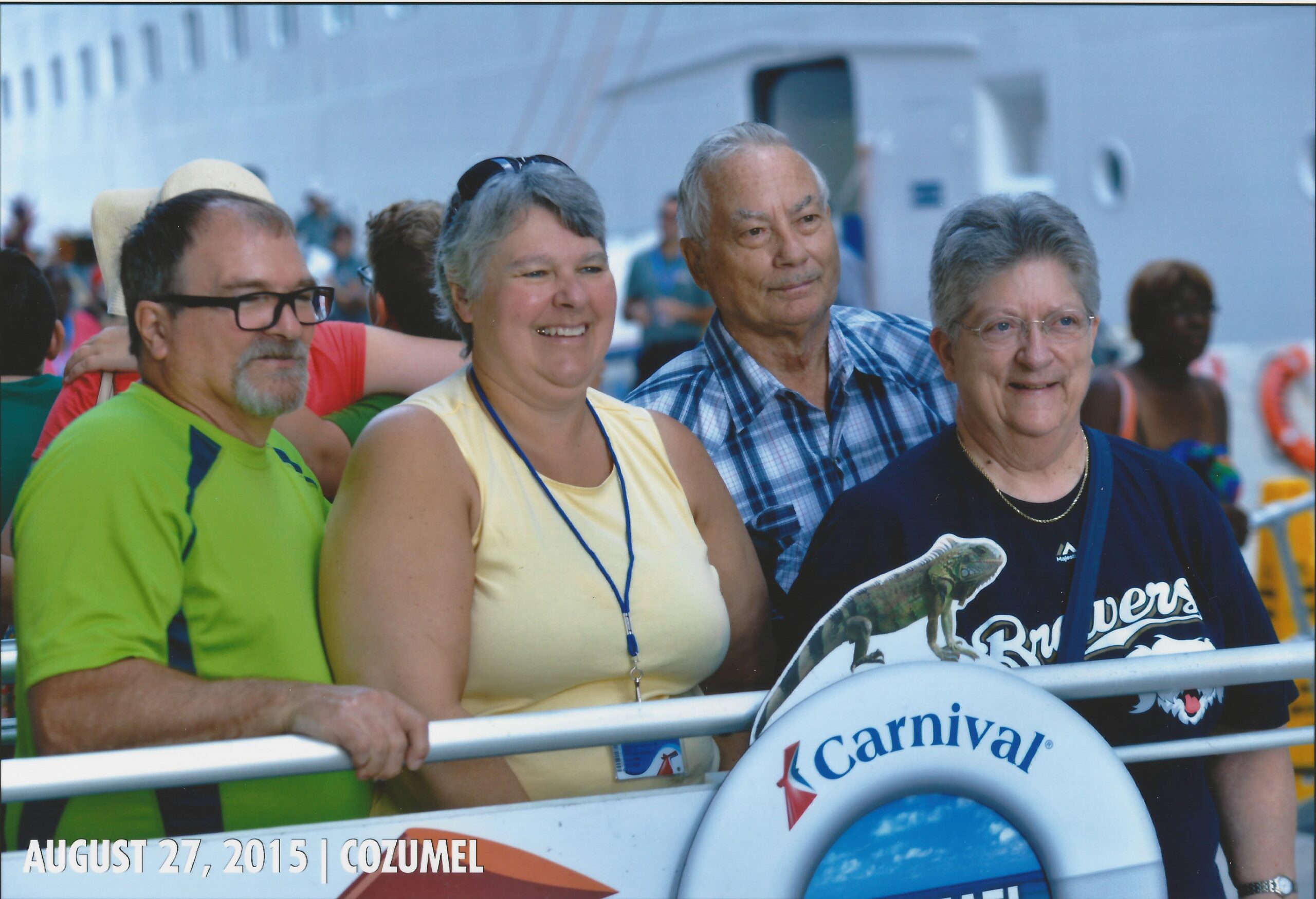 Four adults smile and pose behind a railing at a Carnival cruise event; a Carnival life preserver is visible in the foreground.