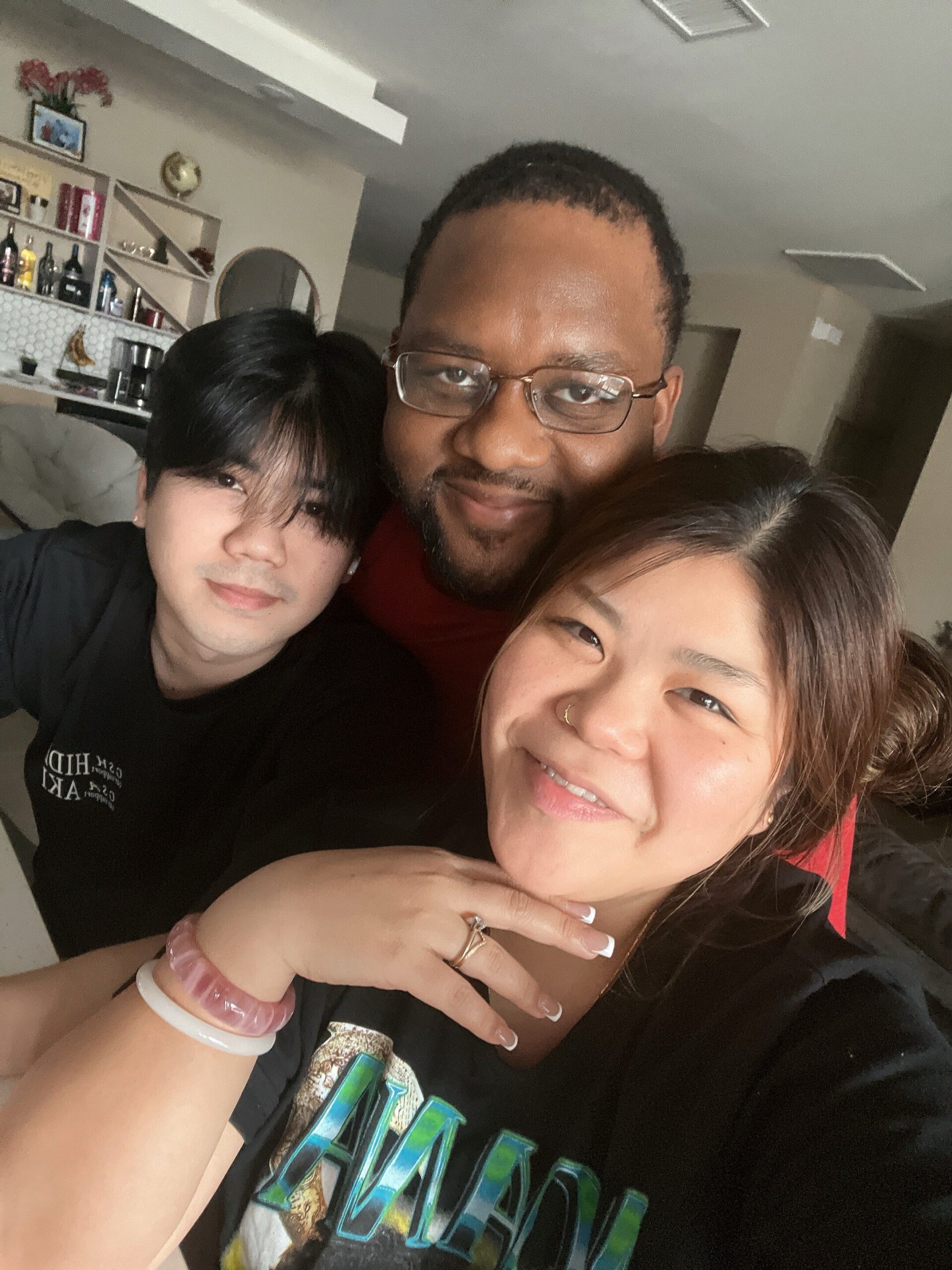 Three friends taking a close, smiling selfie in a living room, with a bar area visible in the background and a woman in the foreground wearing a black shirt and jewelry.