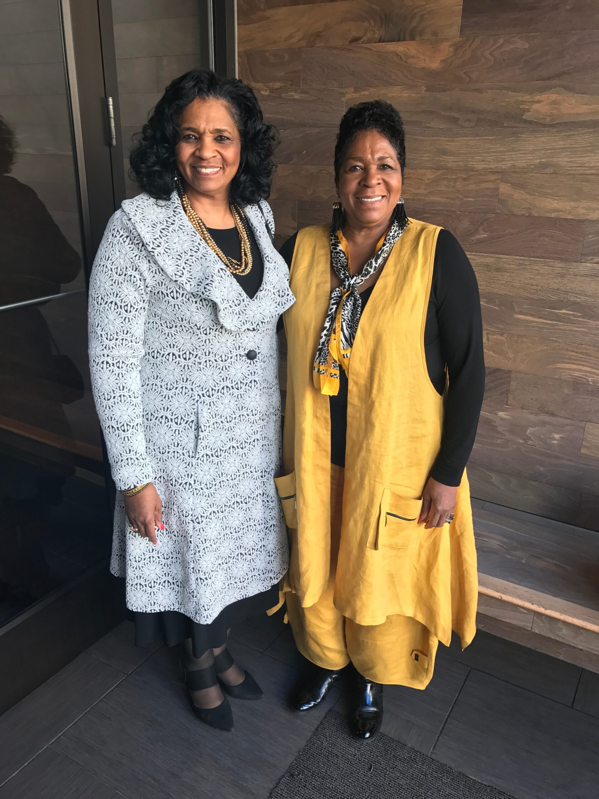 Two smiling women posing together indoors, one wearing a light patterned coat with a black top and gold beads, the other in a mustard vest with a leopard scarf against a wood-paneled wall.