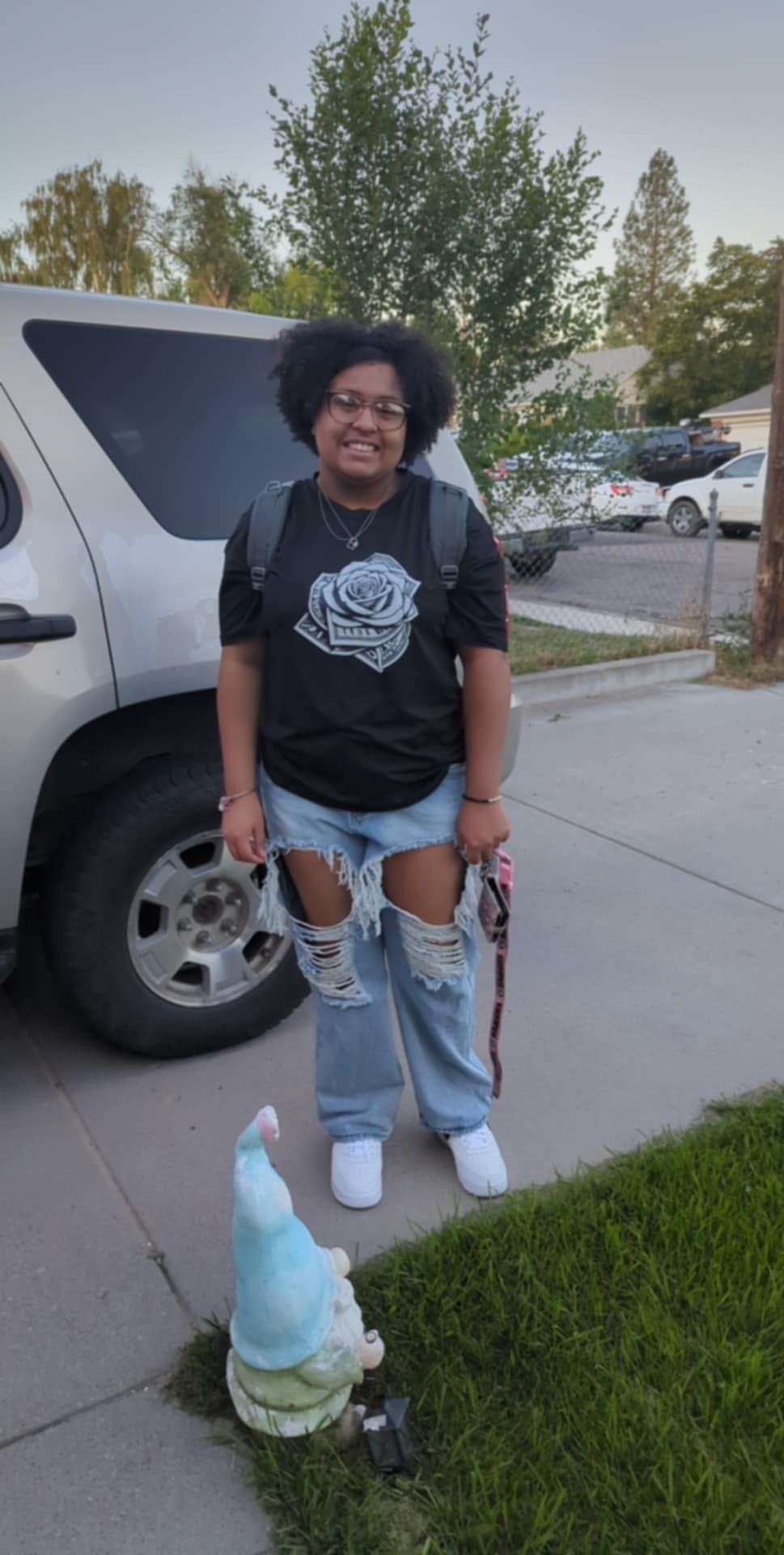 Young woman with backpack standing beside a white van on a suburban street, wearing a black rose-graphic shirt and ripped jeans.
