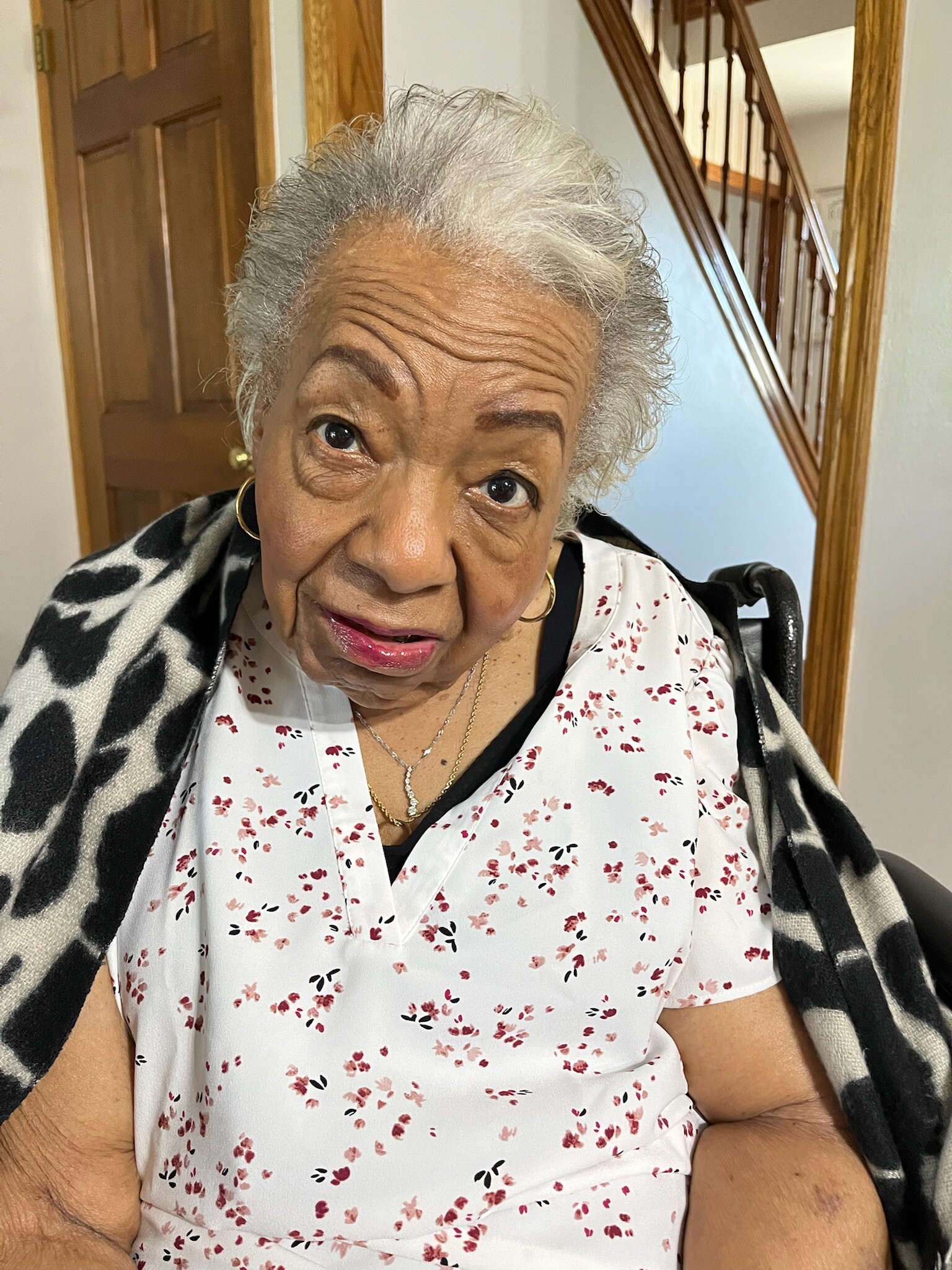Close-up of an elderly woman with gray hair and hoop earrings, wearing a white floral blouse and a black-and-white shawl indoors by a staircase.