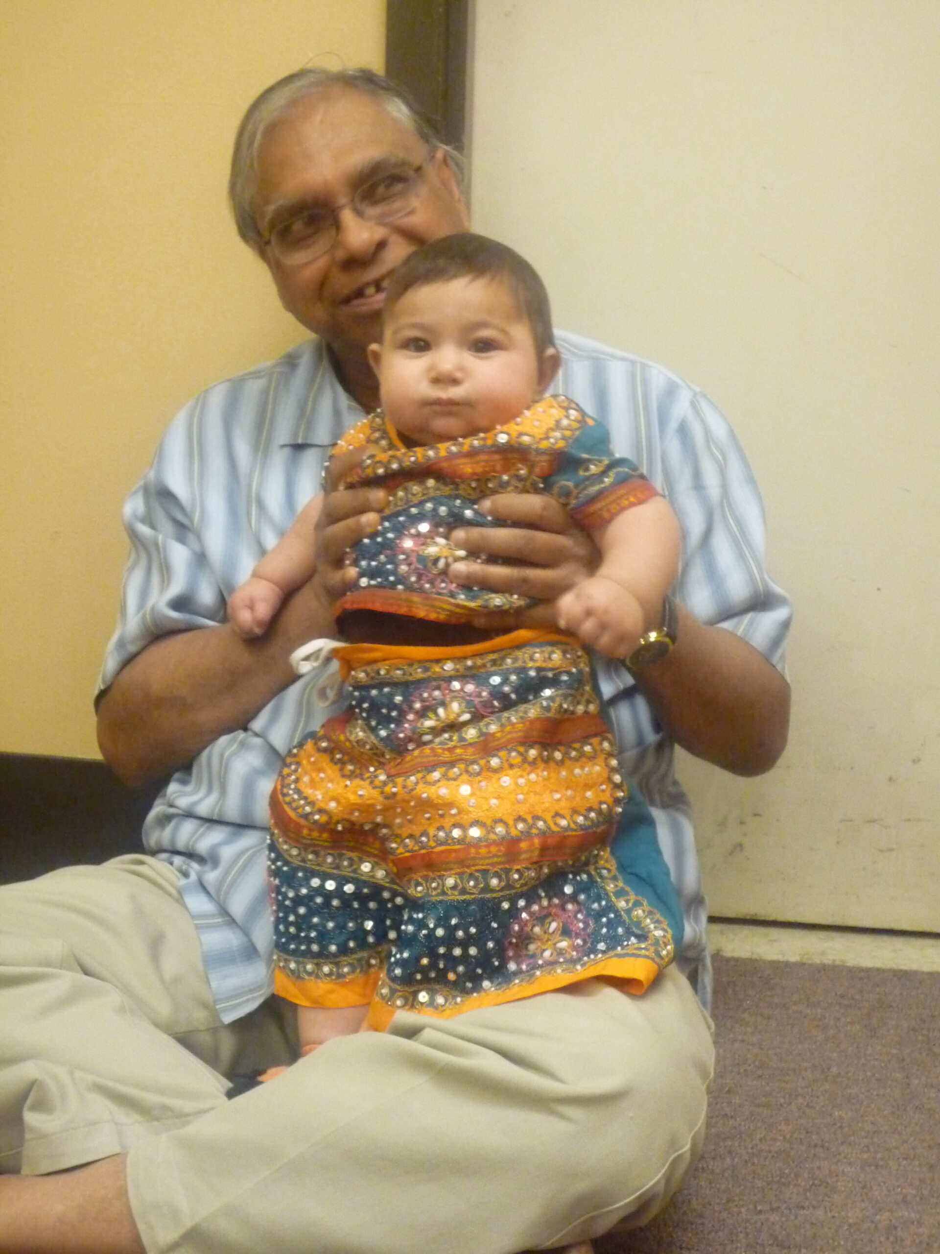 An older man in a striped shirt holds a baby in a bright, embroidered dress, both looking toward the camera in a neutral room.