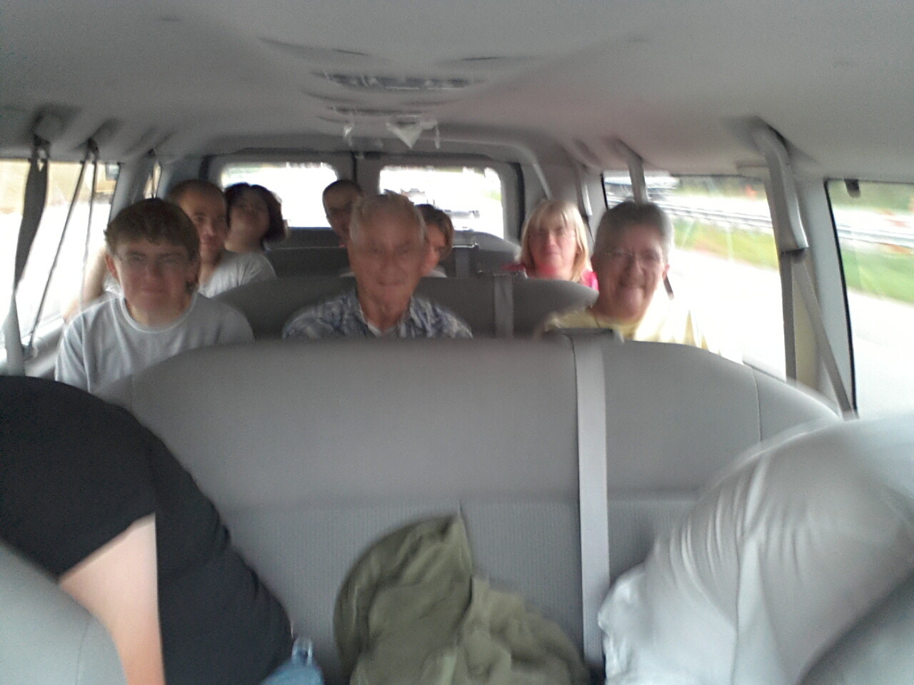 A group of people sitting in a moving van, facing the camera and smiling from the back rows.