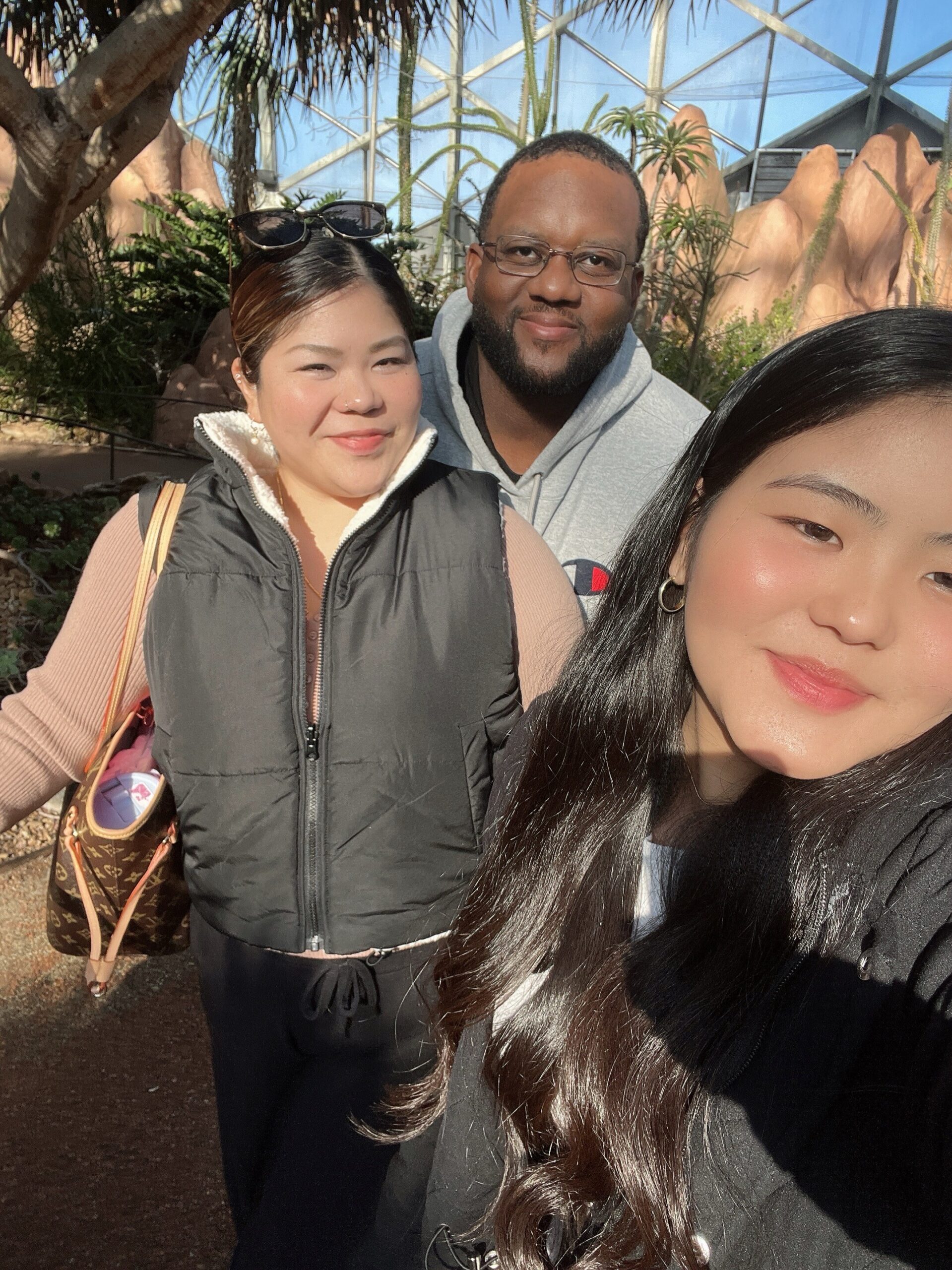 Three friends pose for a sunny selfie in a glass greenhouse with tropical plants in the background.