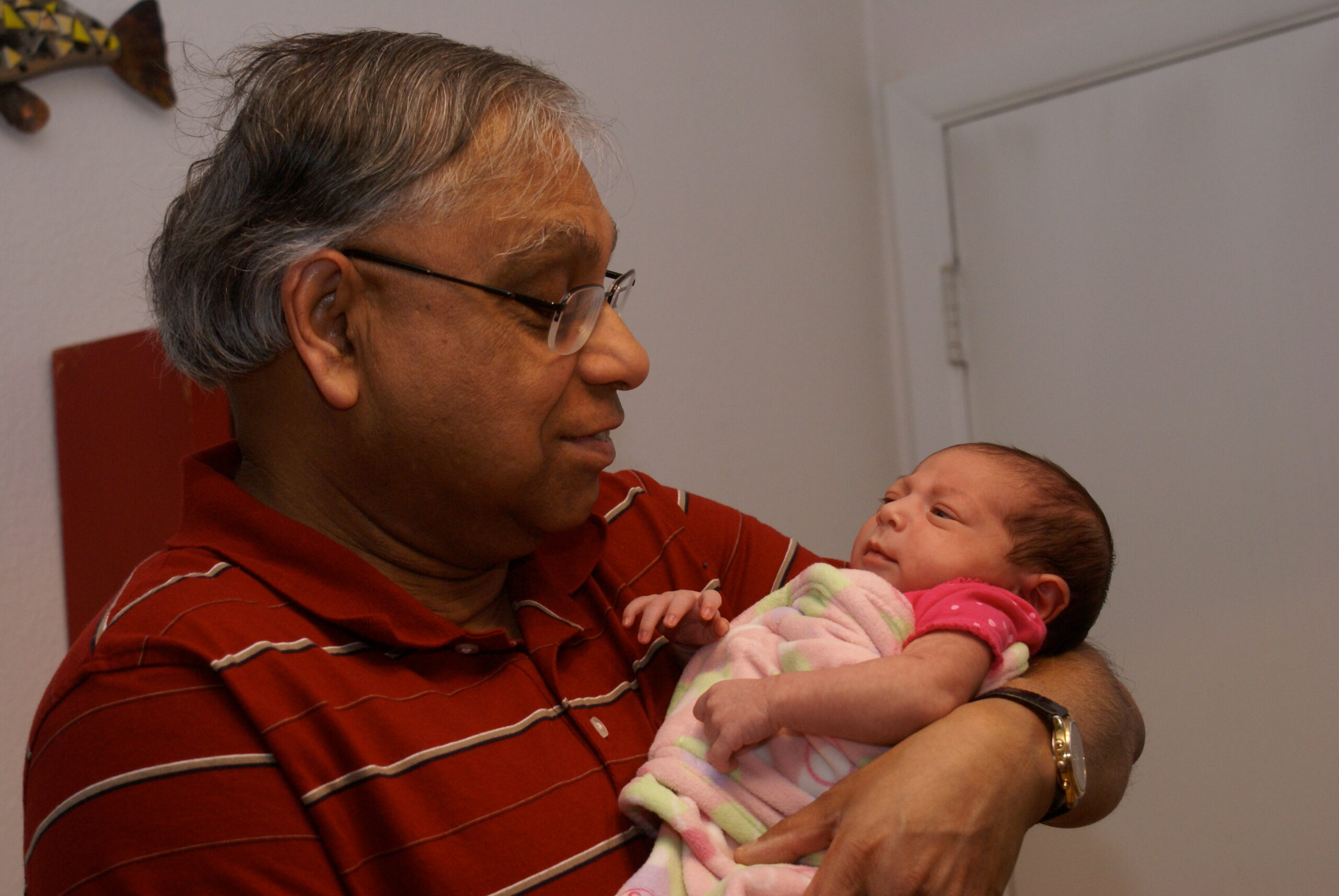 Older man with glasses holding a newborn baby in a pink blanket indoors, smiling at the infant.