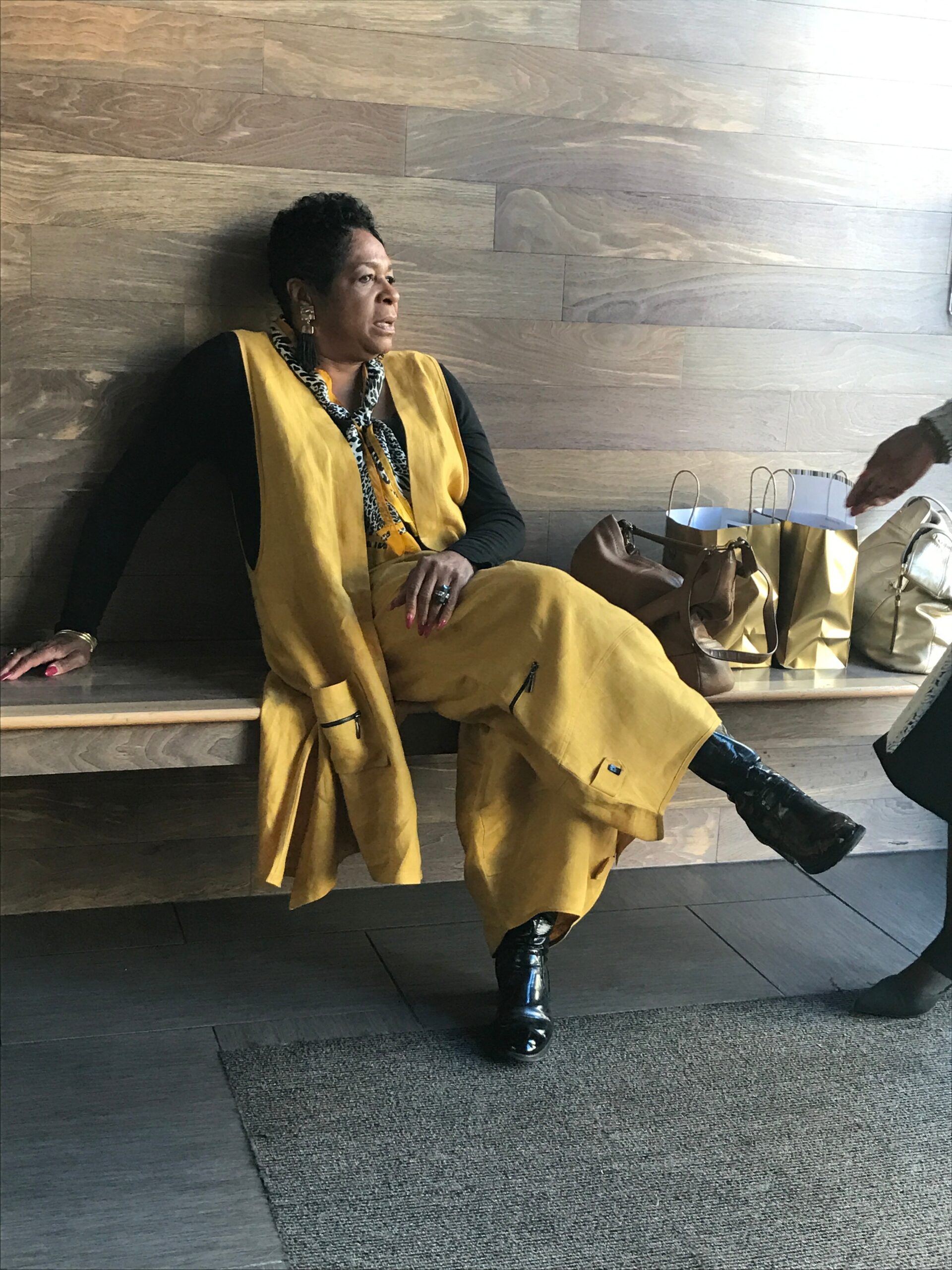 Woman in a mustard yellow coat and scarf sits on a wooden bench, with shopping bags nearby.