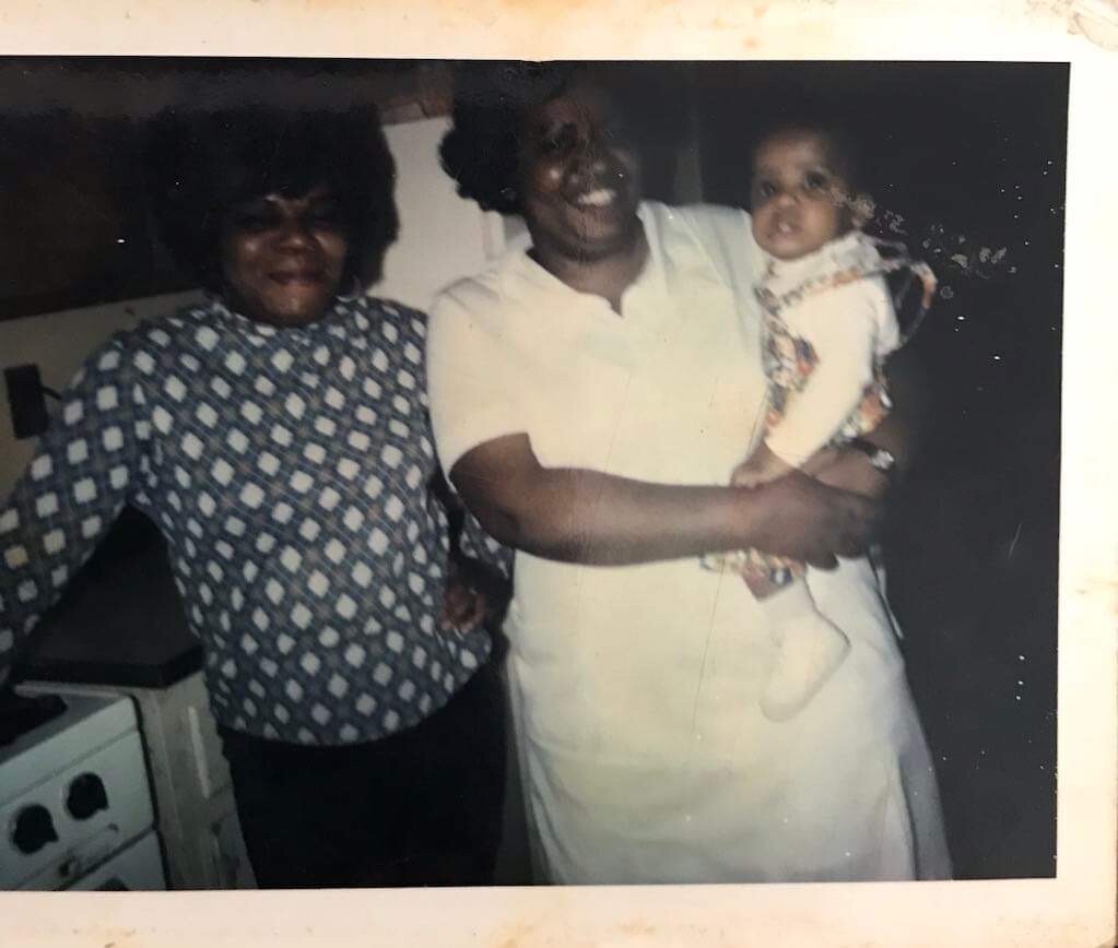 Three generations: a woman in a patterned blouse, a smiling woman in white holding a baby in a kitchen.