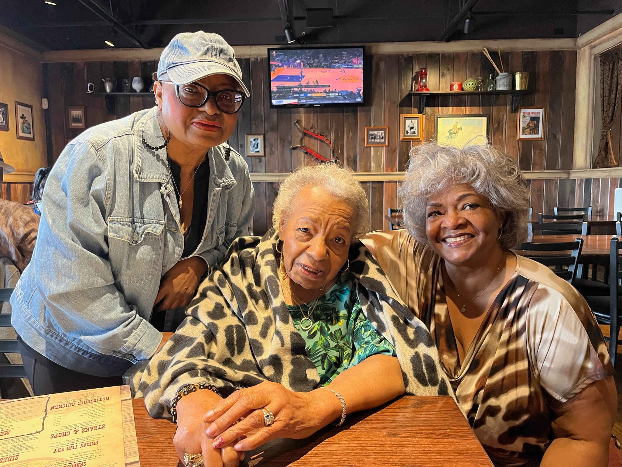 Three smiling senior women pose together at a wood-paneled restaurant table, with a TV and framed art in the background.
