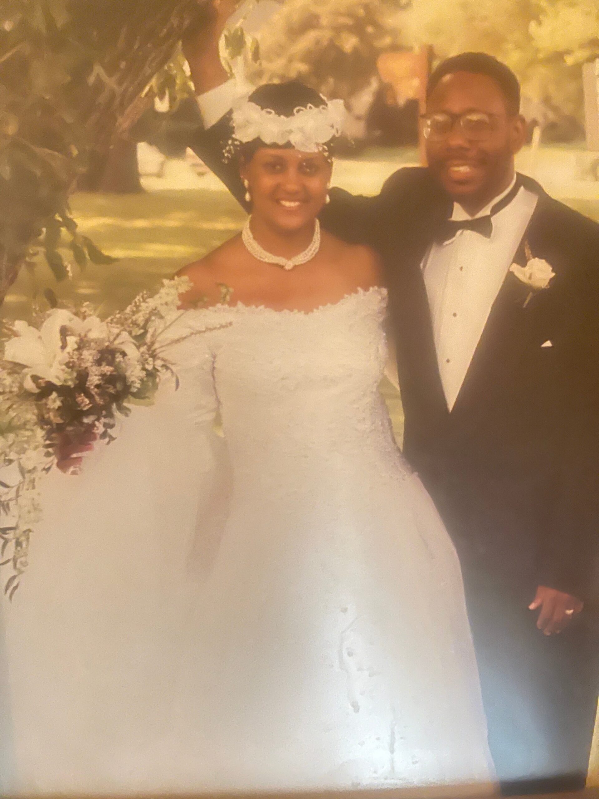 Bride in an off-shoulder white wedding gown with a floral headpiece and pearl necklace stands next to a smiling groom in a black tuxedo outdoors, holding a bouquet of flowers.