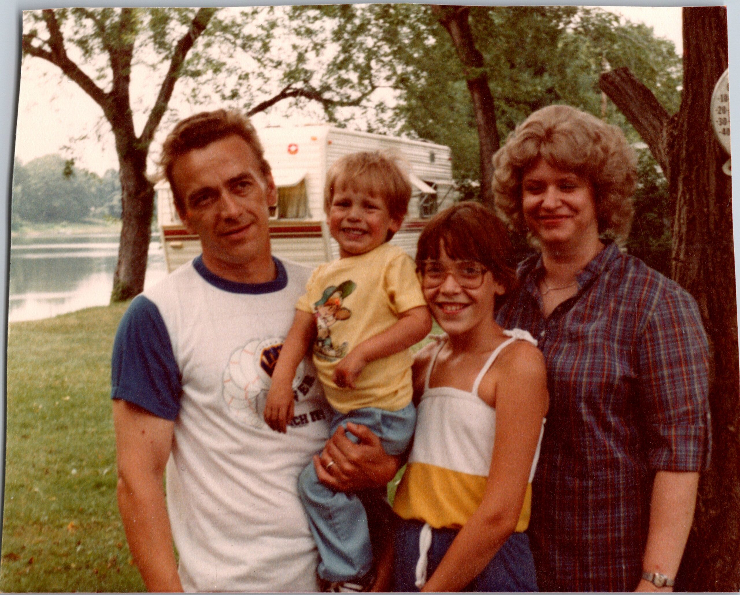 Group photo of a man, a smiling child, and a woman outdoors near a trailer in a park setting.