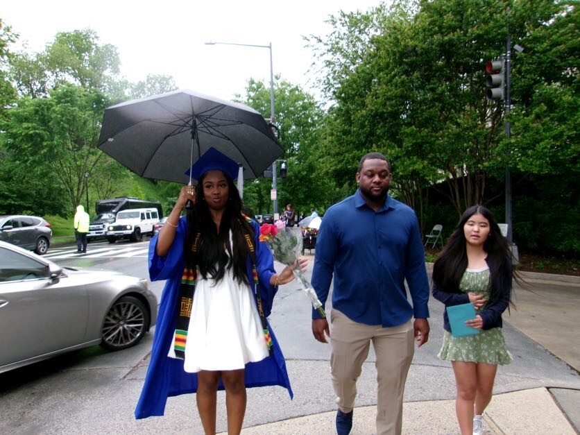 Graduate in blue cap and gown holding an umbrella walks with two companions on a rainy street after a ceremony, a man in a blue shirt and a girl with a folder beside her