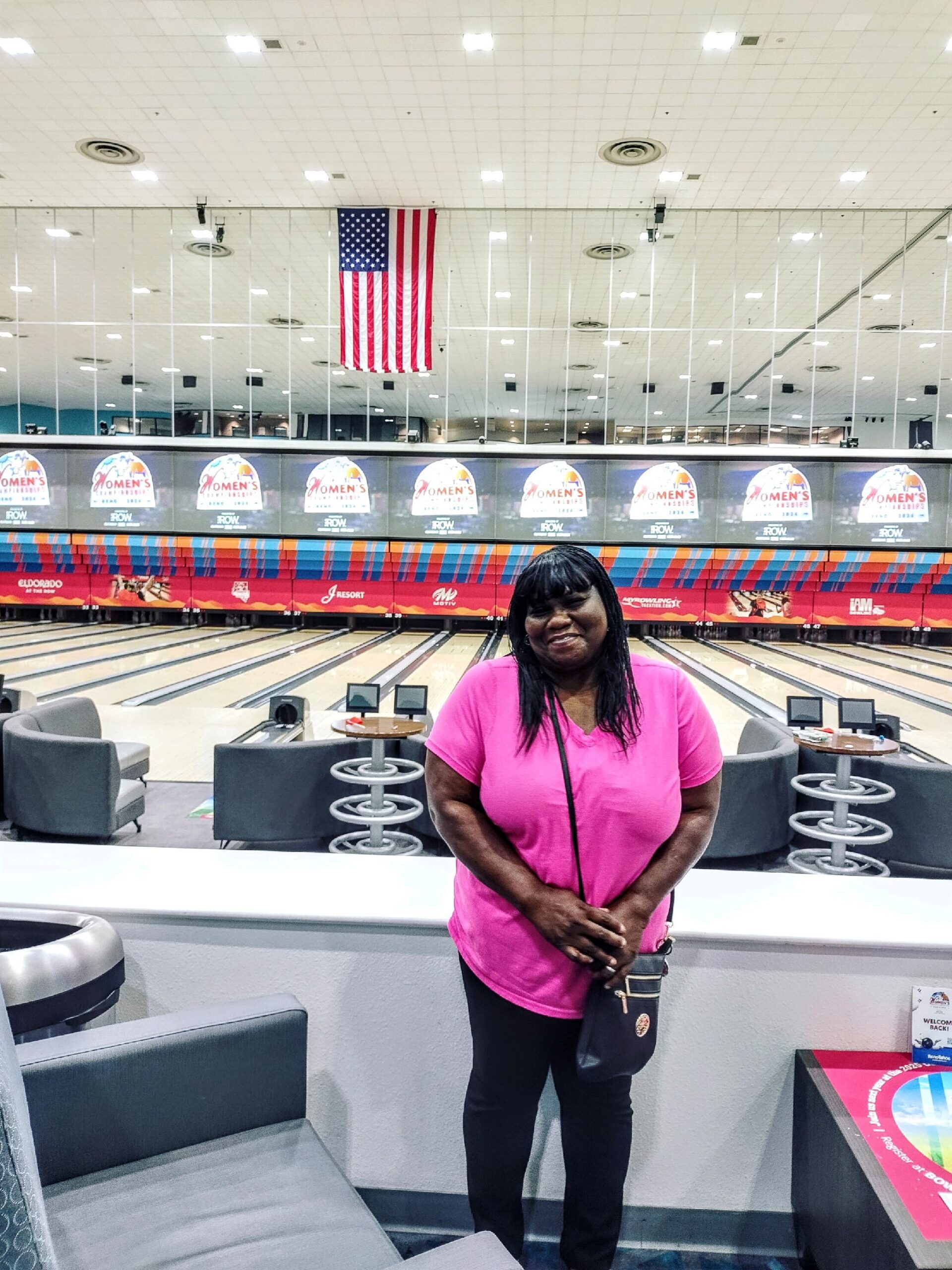 Smiling woman in a pink shirt stands at the edge of a bowling alley, with lanes, seating, and an American flag overhead.