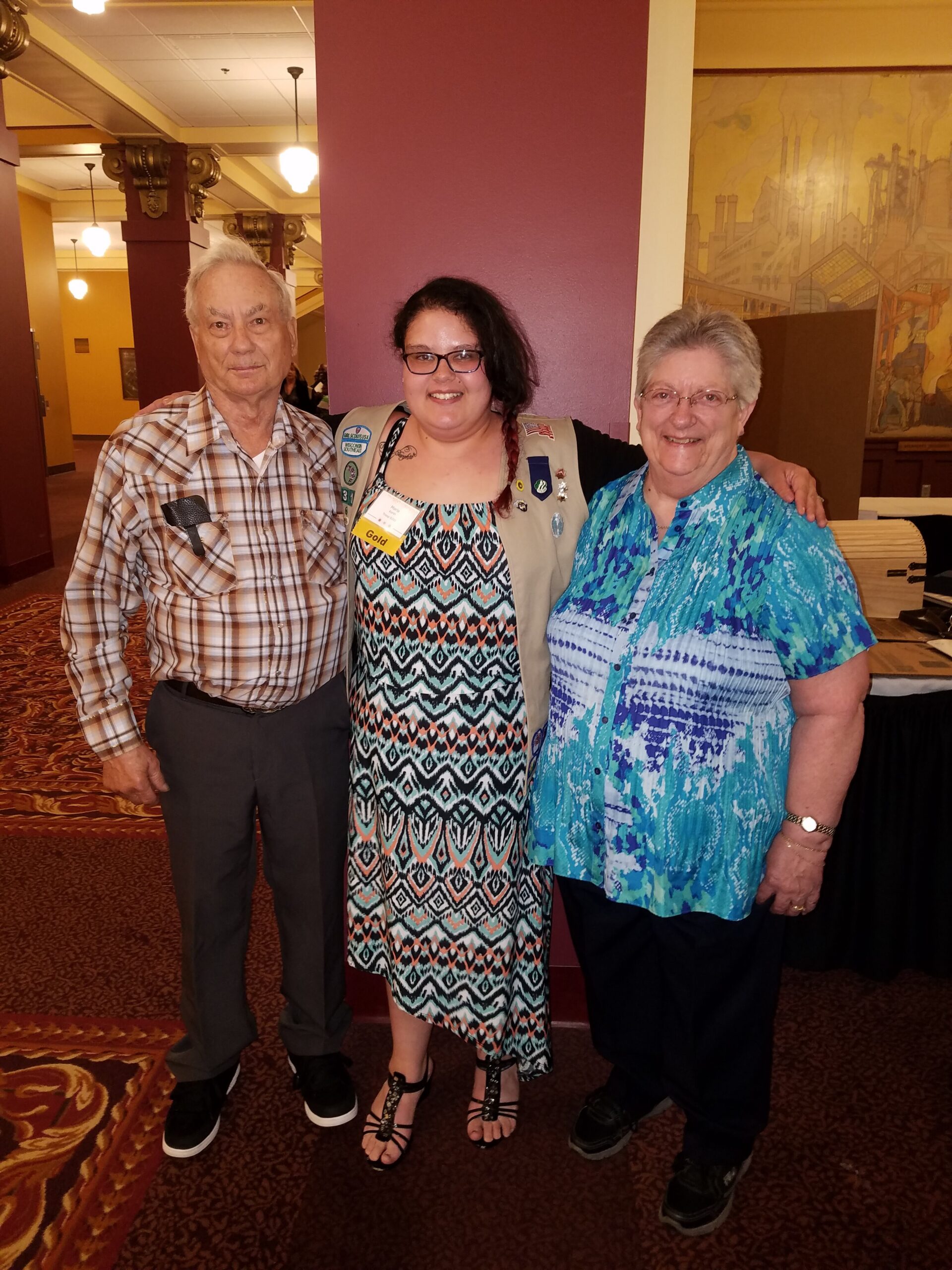 Three adults posed together indoors, smiling for a group photo: an older man in a plaid shirt on the left, a woman with badges in the middle, and a woman in a blue patterned top on the right.