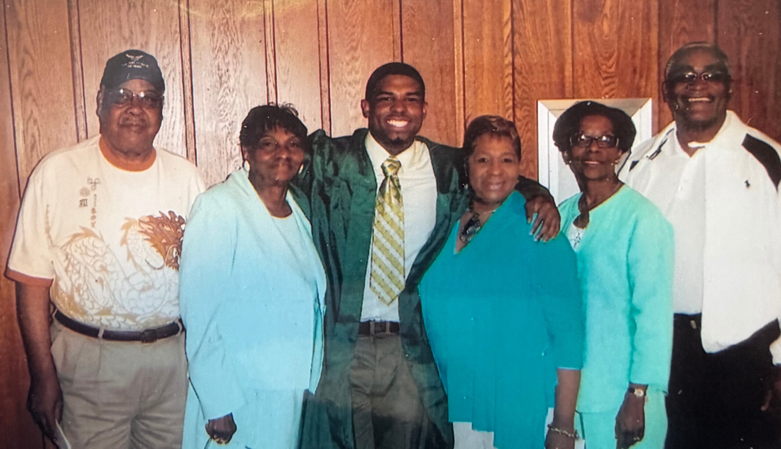 Six adults posing indoors in front of wood paneling; center man wears a green gown and striped yellow tie, flanked by others in teal outfits and casual clothes, all smiling.