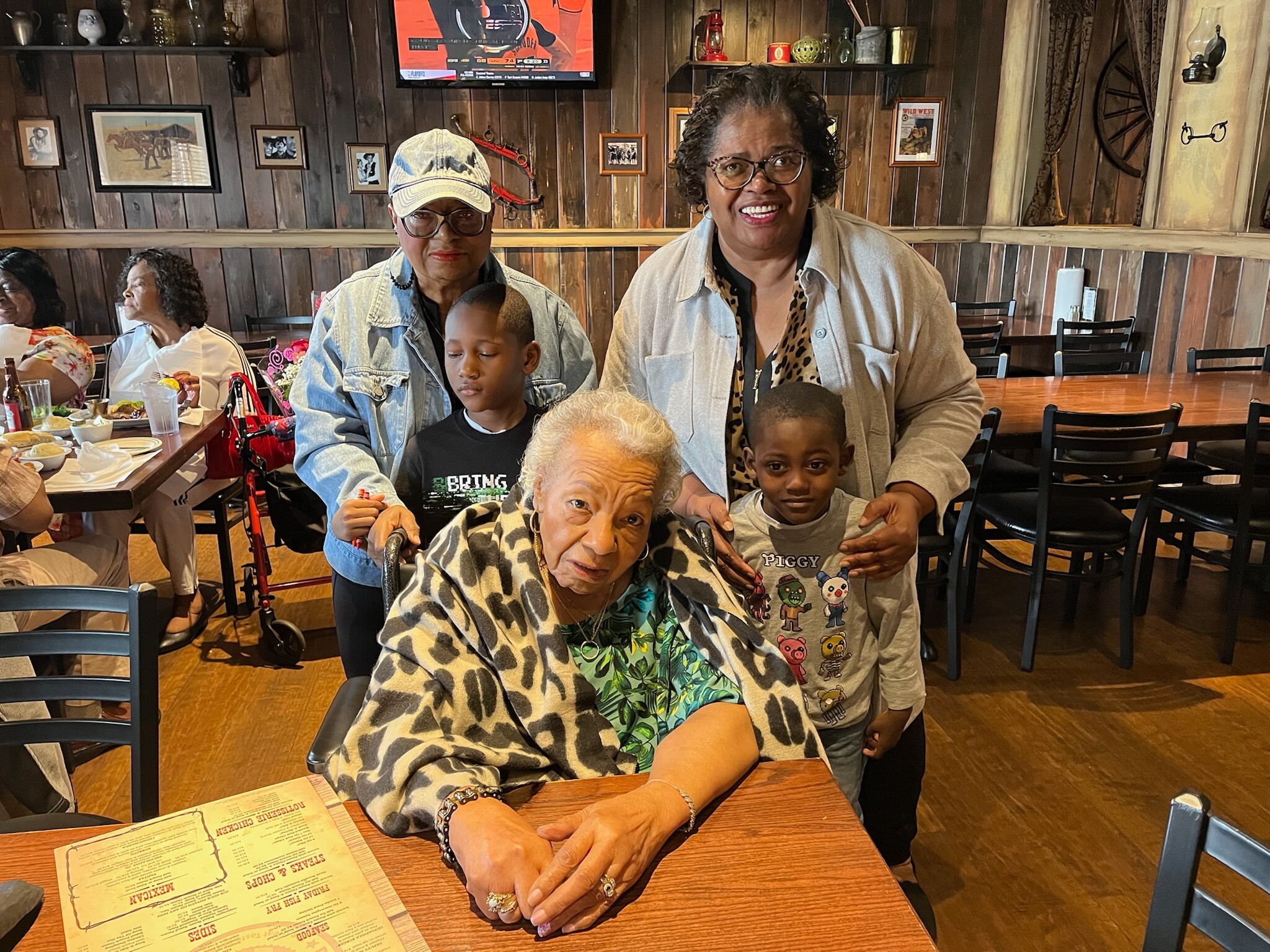 Smiling group at a rustic restaurant table, with an elderly woman in a wheelchair in the foreground wearing a leopard print shawl and a man and child beside her. Behind them, two women stand and chat, and other diners sit at tables in a wood-paneled room.