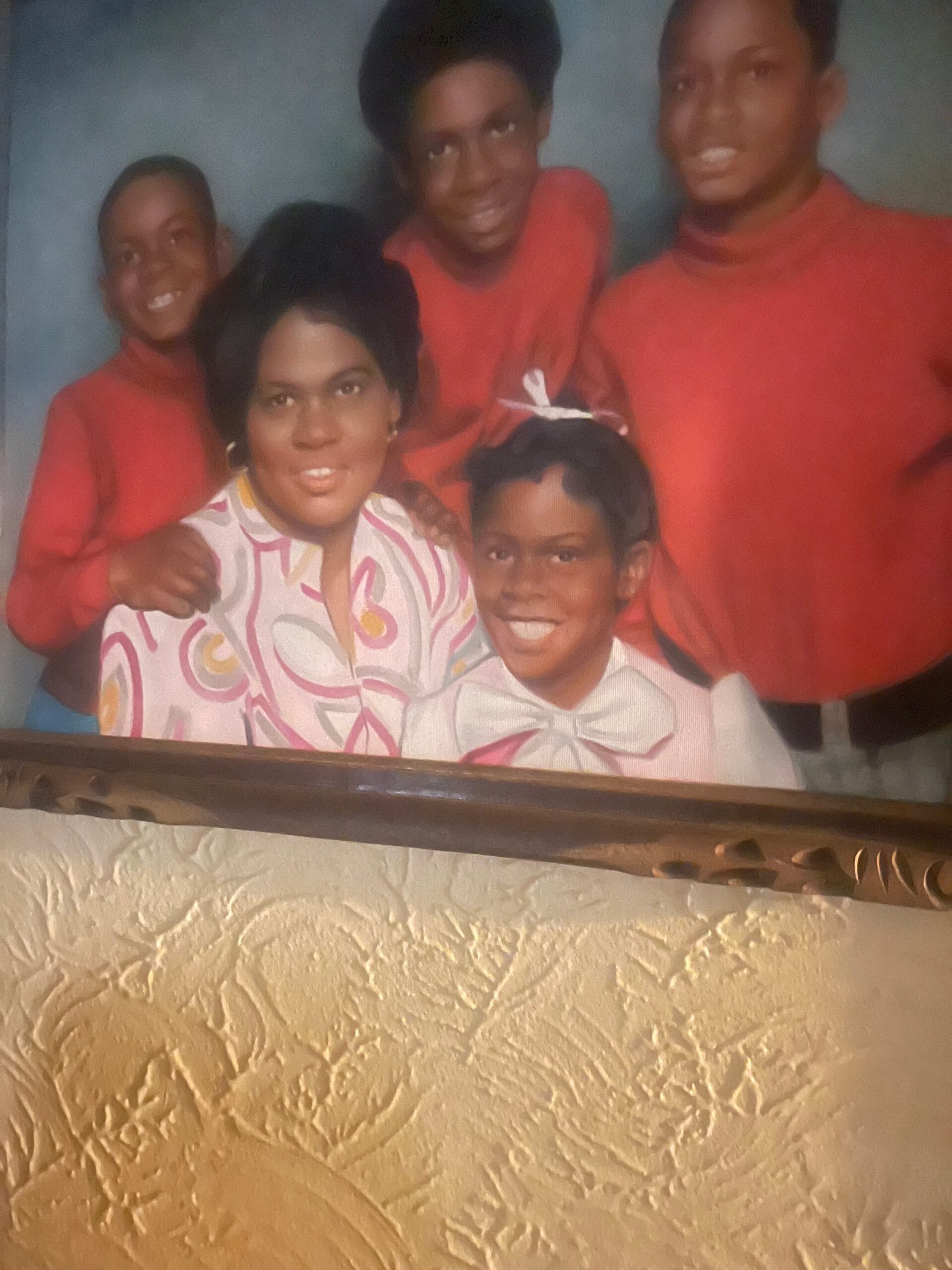 Group of five people posing closely together indoors, smiling, with most wearing red tops; a smiling woman in a patterned blouse sits in front with two children beside her and others around her.