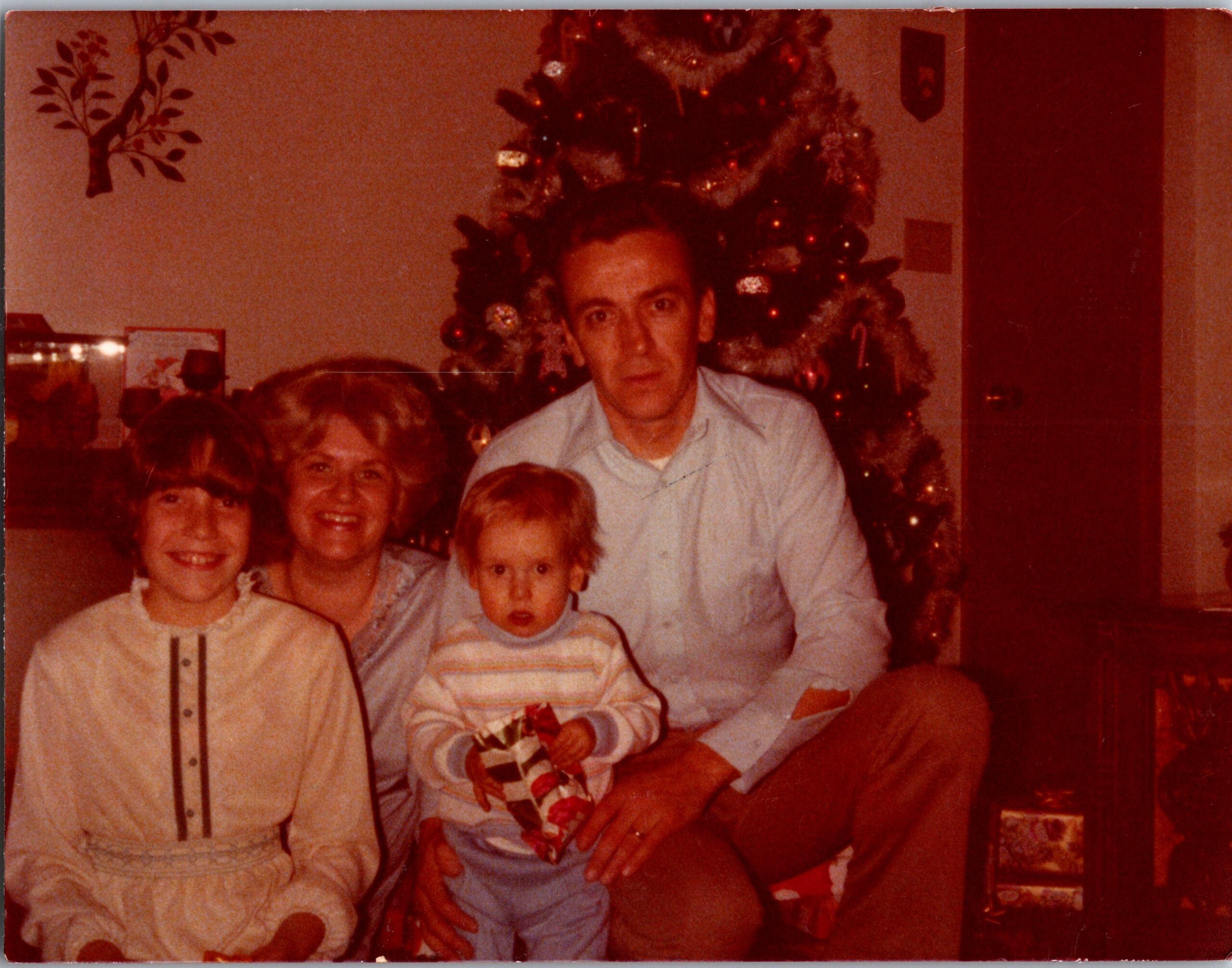 A family poses indoors around a decorated Christmas tree with wrapped gifts nearby.