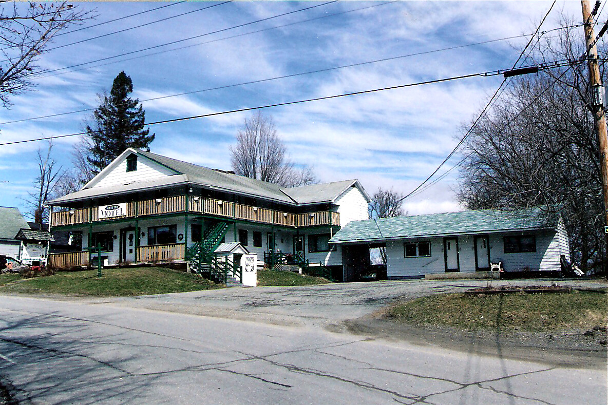 Two-story motel with a wooden balcony and green stairwell beside a white, single-story annex on a rural road. Inset sign reads MOTEL.
