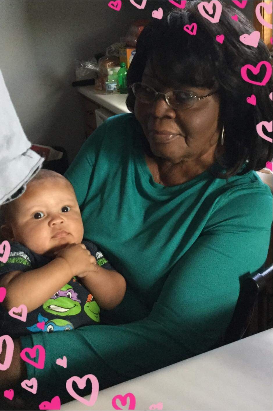 Grandmother in a teal shirt holding a baby in a kitchen, with a pink heart border around the image.