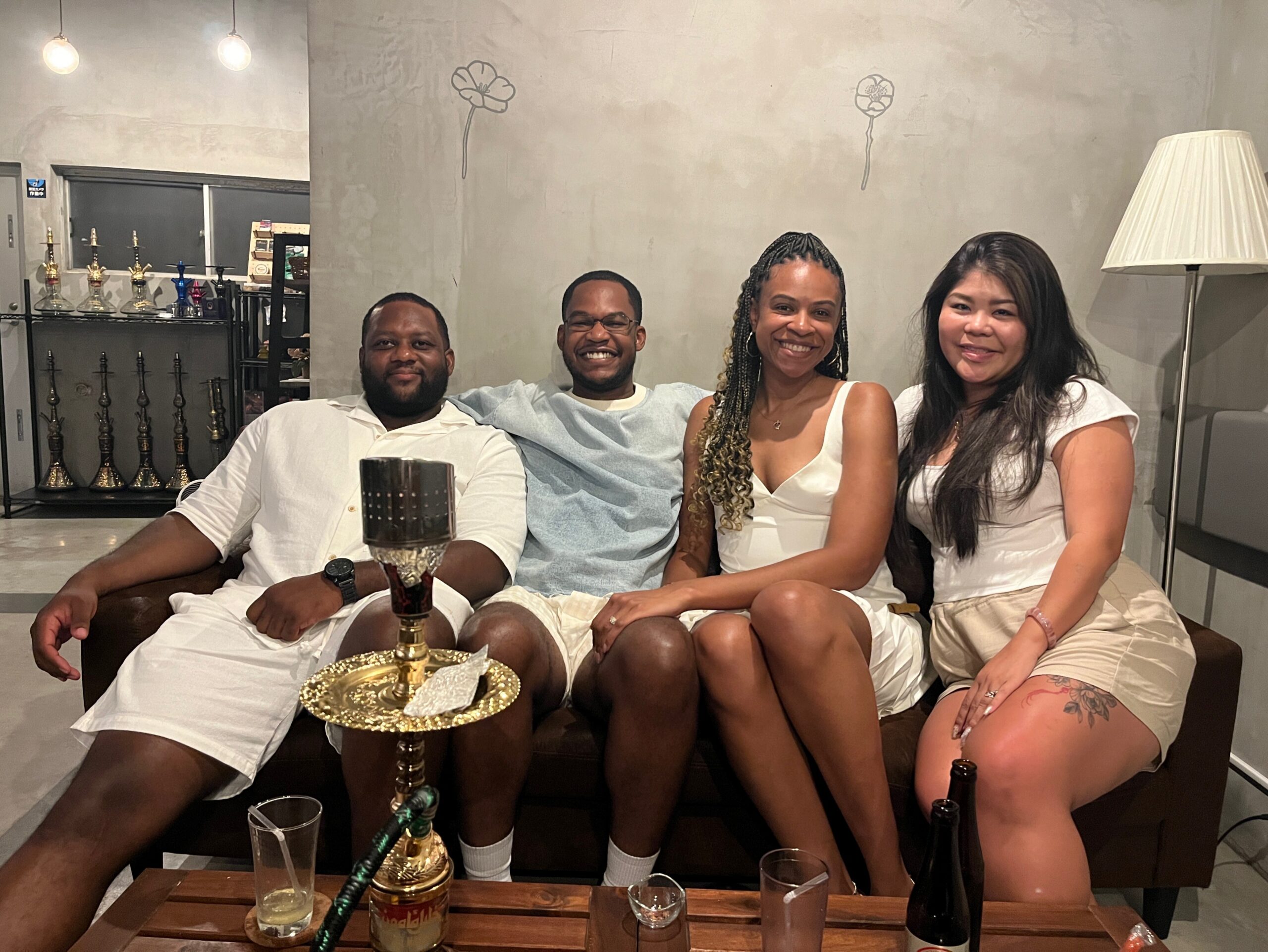 Four friends sitting on a couch indoors, smiling at the camera, with a hookah and drinks on a wooden table in the foreground and warm lighting in a lounge-like room behind them.