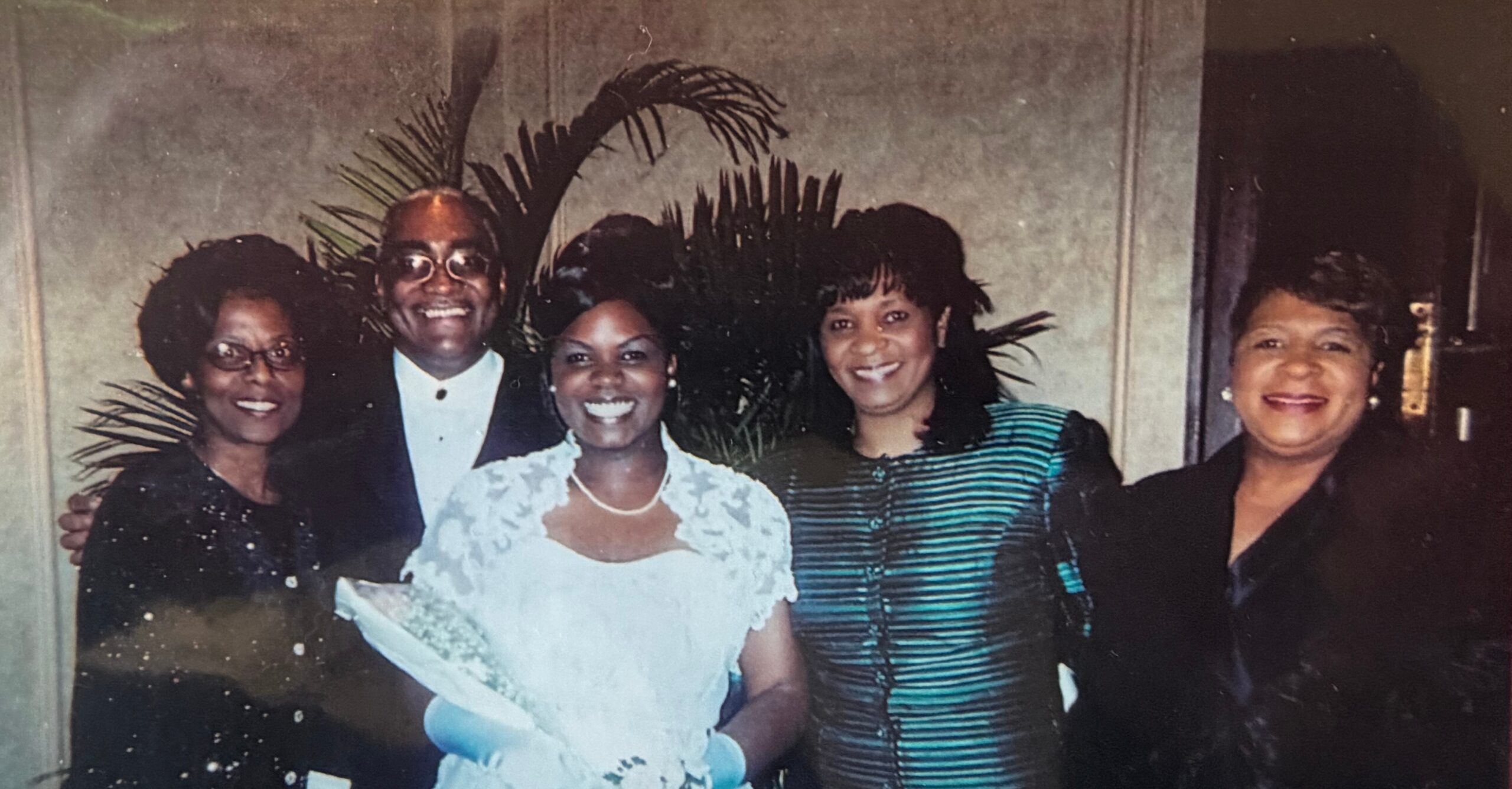 Bride in a white lace wedding gown holds a bouquet and stands with four smiling guests in formal attire at a party.