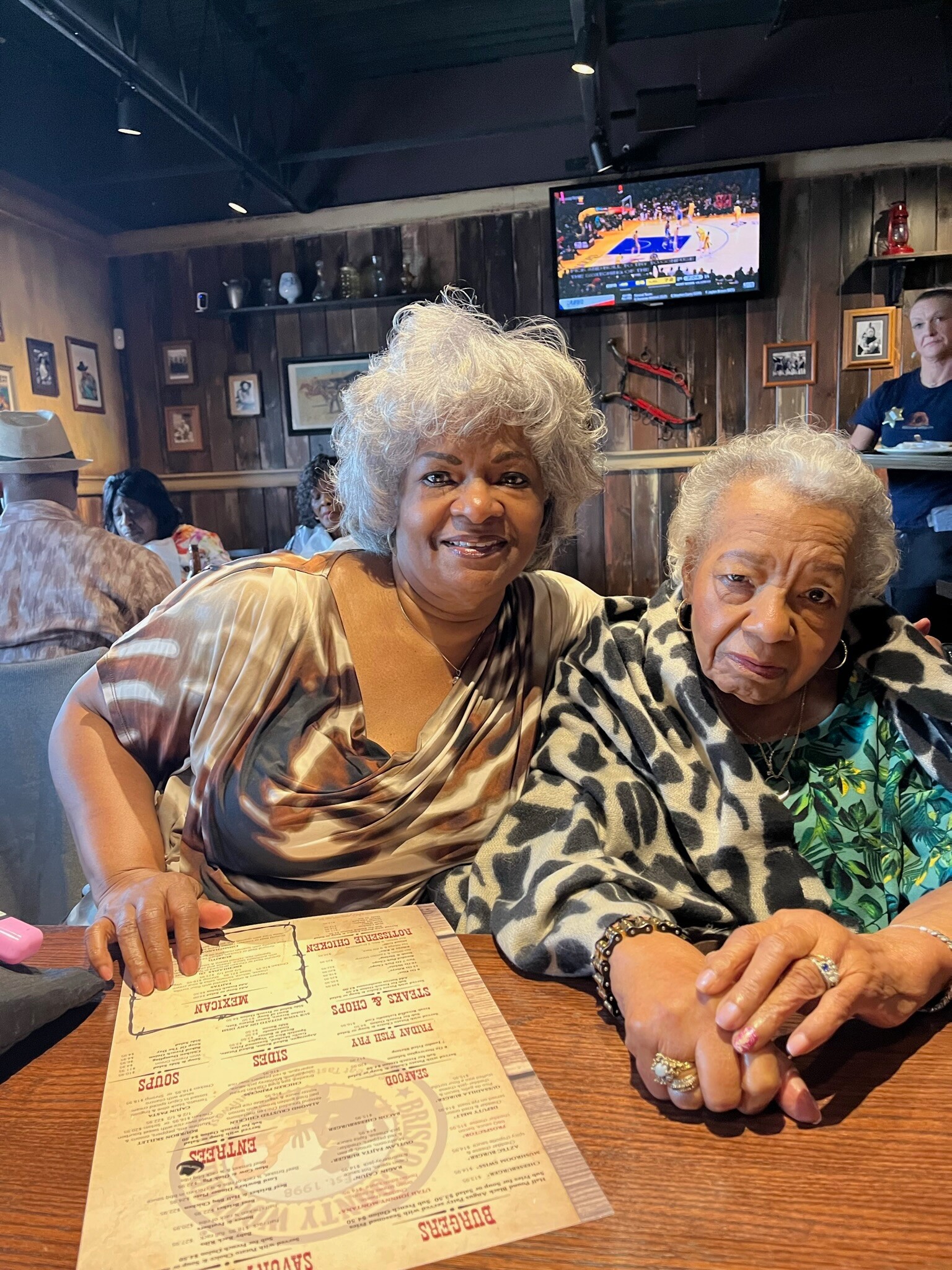 Two smiling elderly women sit at a wooden table in a restaurant, hands resting on a menu, with a TV playing basketball in the background and warm wood-paneled walls.