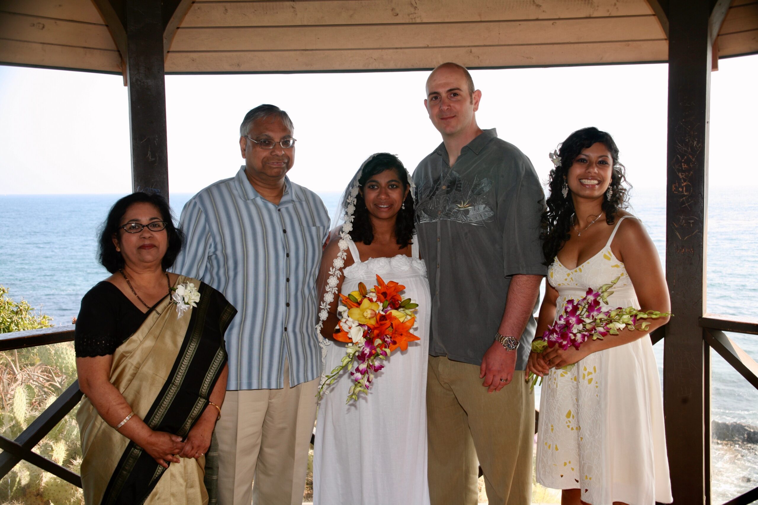 Group of five people posing on a seaside balcony, two brides wearing white dresses and bouquets, with family members on either side, ocean in background