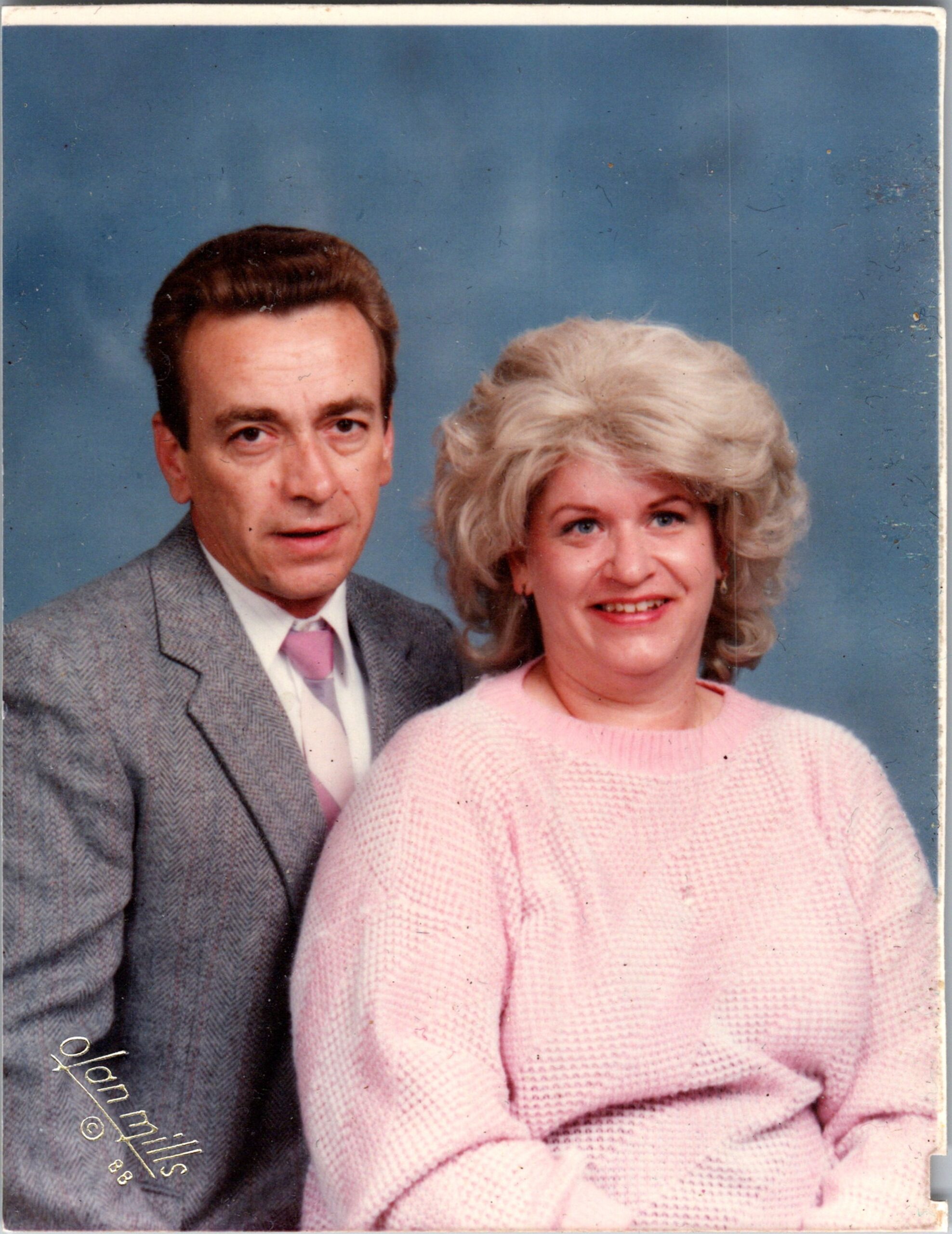 Older couple posed for a studio portrait; man in a gray suit and pink tie stands beside a woman in a pink sweater against a blue backdrop.