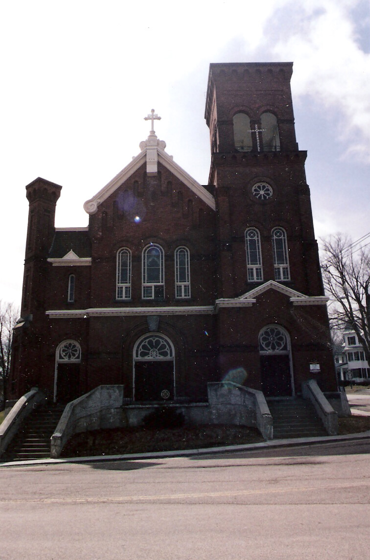 Brick church with a tall square bell tower, arched windows, and a cross on the gable; stairs lead to the entrances.