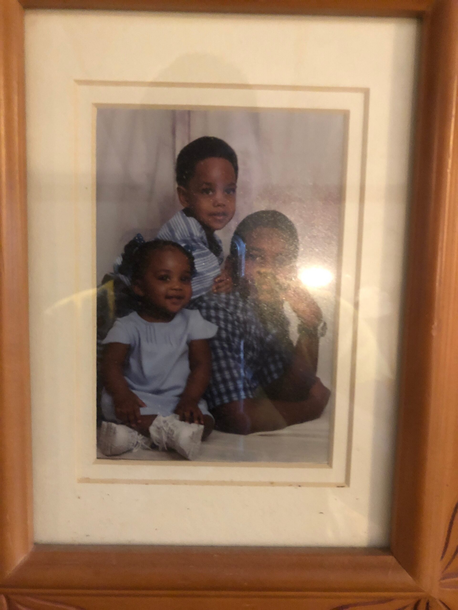 Framed family portrait of three young children posed together, with a boy in a blue striped shirt in the center and a girl on the left wearing a bow.