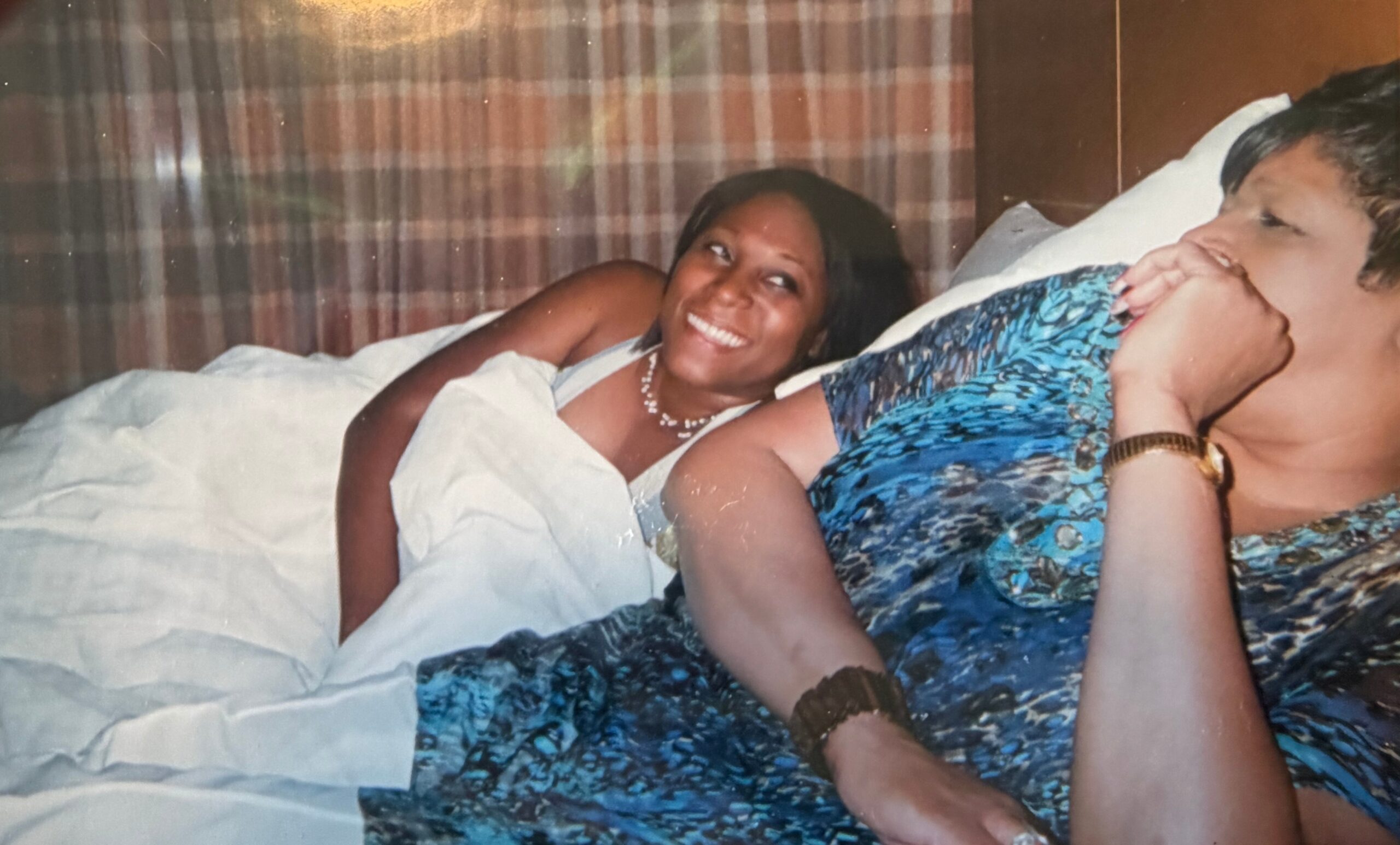 Two people lying together in bed, smiling at the camera; the woman wears a white outfit and necklace, the man rests nearby in a blue patterned shirt.