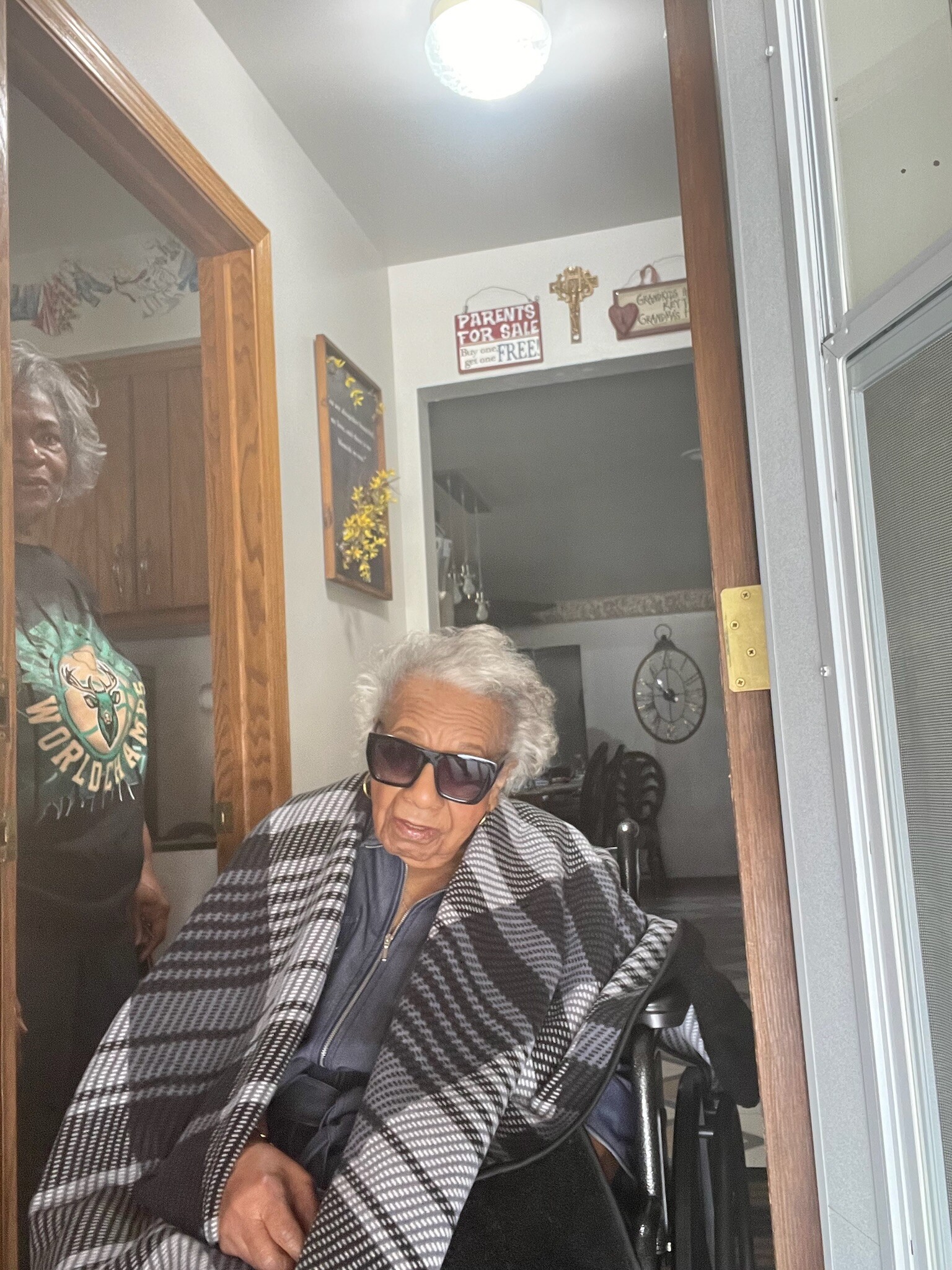 Elderly woman in a wheelchair wearing a black-and-white checkered shawl sits at a doorway inside a home, with another person standing nearby in a green shirt.