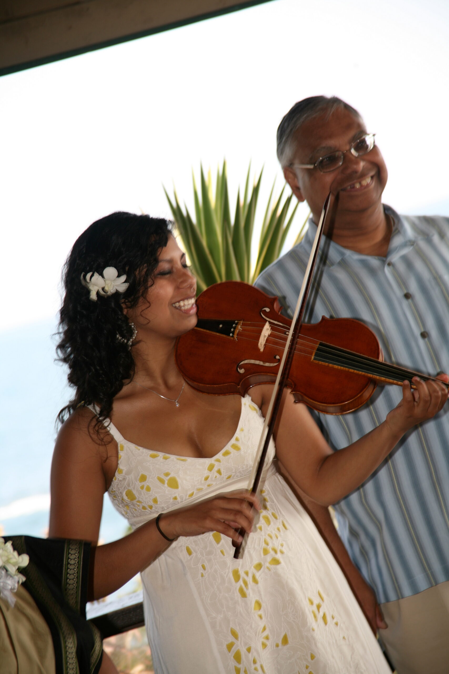 Smiling woman in a white dress with a white flower in her hair plays a violin outdoors, with a man in a striped shirt beside her.