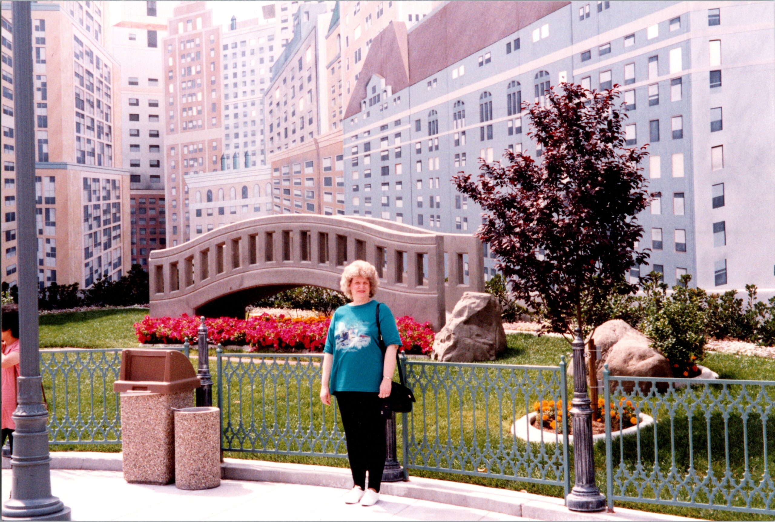 Older woman in a turquoise top stands in a city courtyard with a curved stone bridge, pastel buildings, and flower beds behind a decorative green fence.