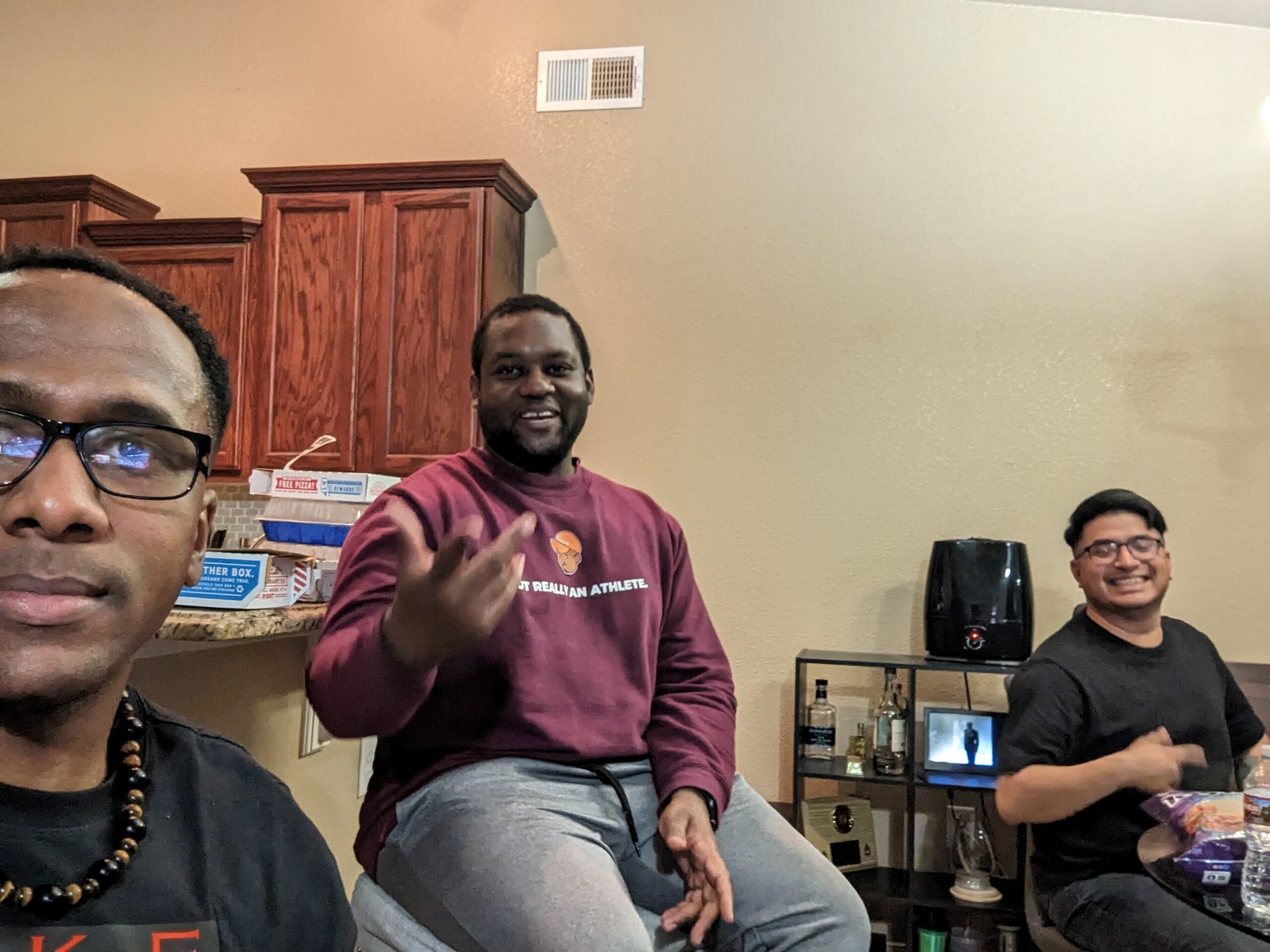 Three men pose for a casual selfie in a home kitchen/living area, smiling with cabinets and a beverage station in the background.