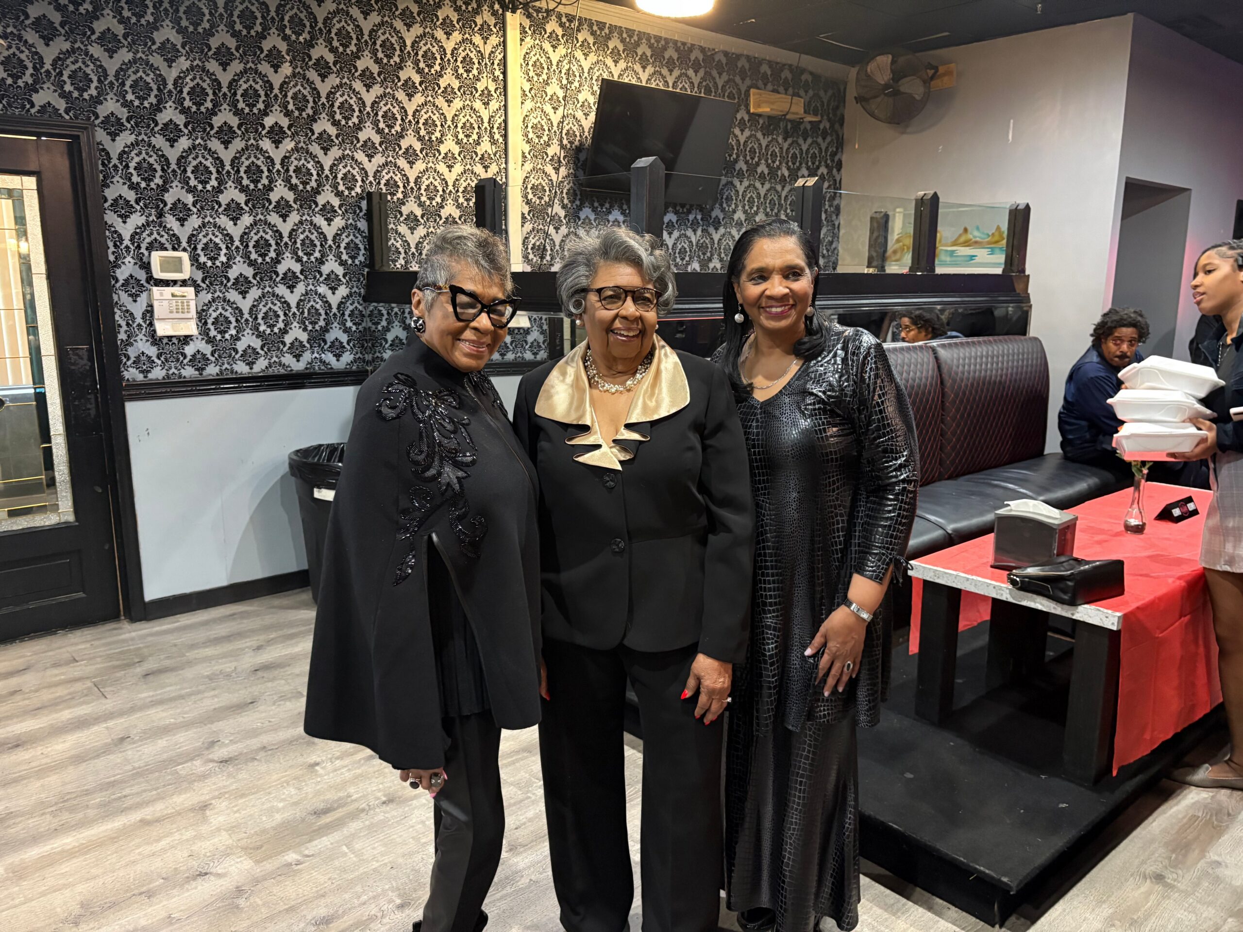 Three women in black formal outfits pose together indoors against a damask-patterned wall, smiling for the photo.