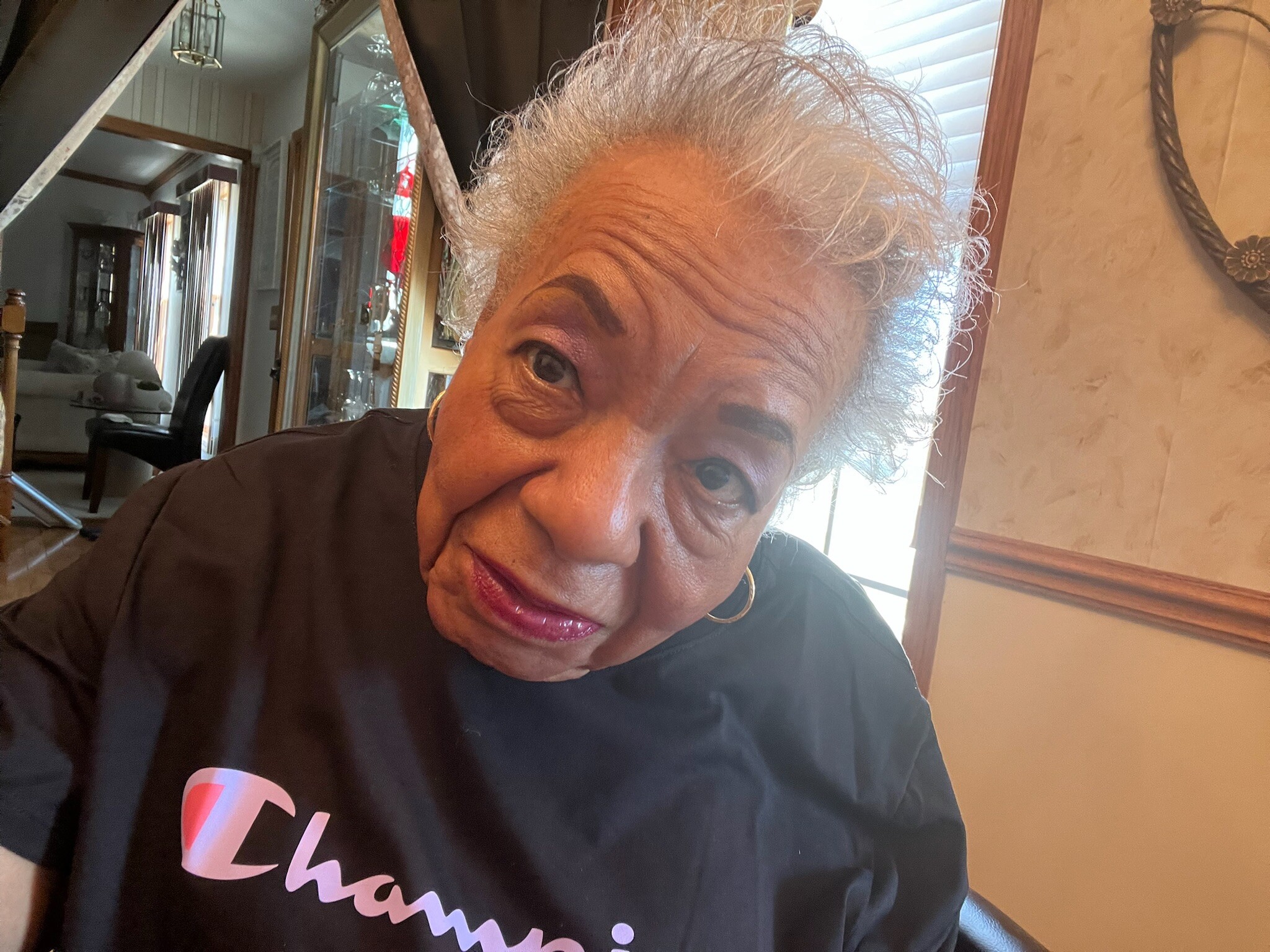 Close-up of an elderly woman with gray hair and pink lipstick, wearing a black shirt with pink script text, indoors.