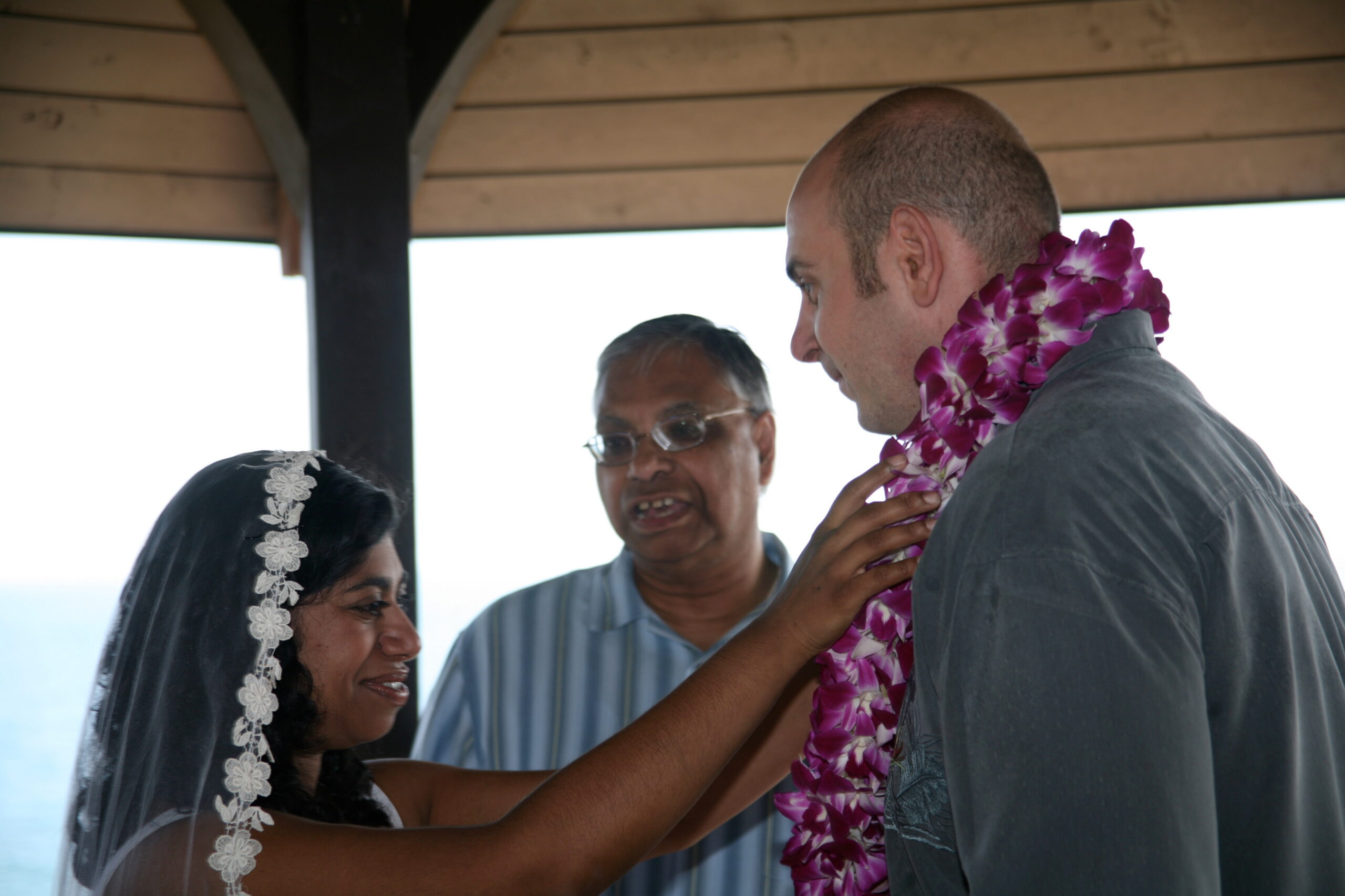 Bride in a lace veil and a man in a dark shirt share a moment as guests help adjust a pink lei; ocean and a wooden structure in the background.
