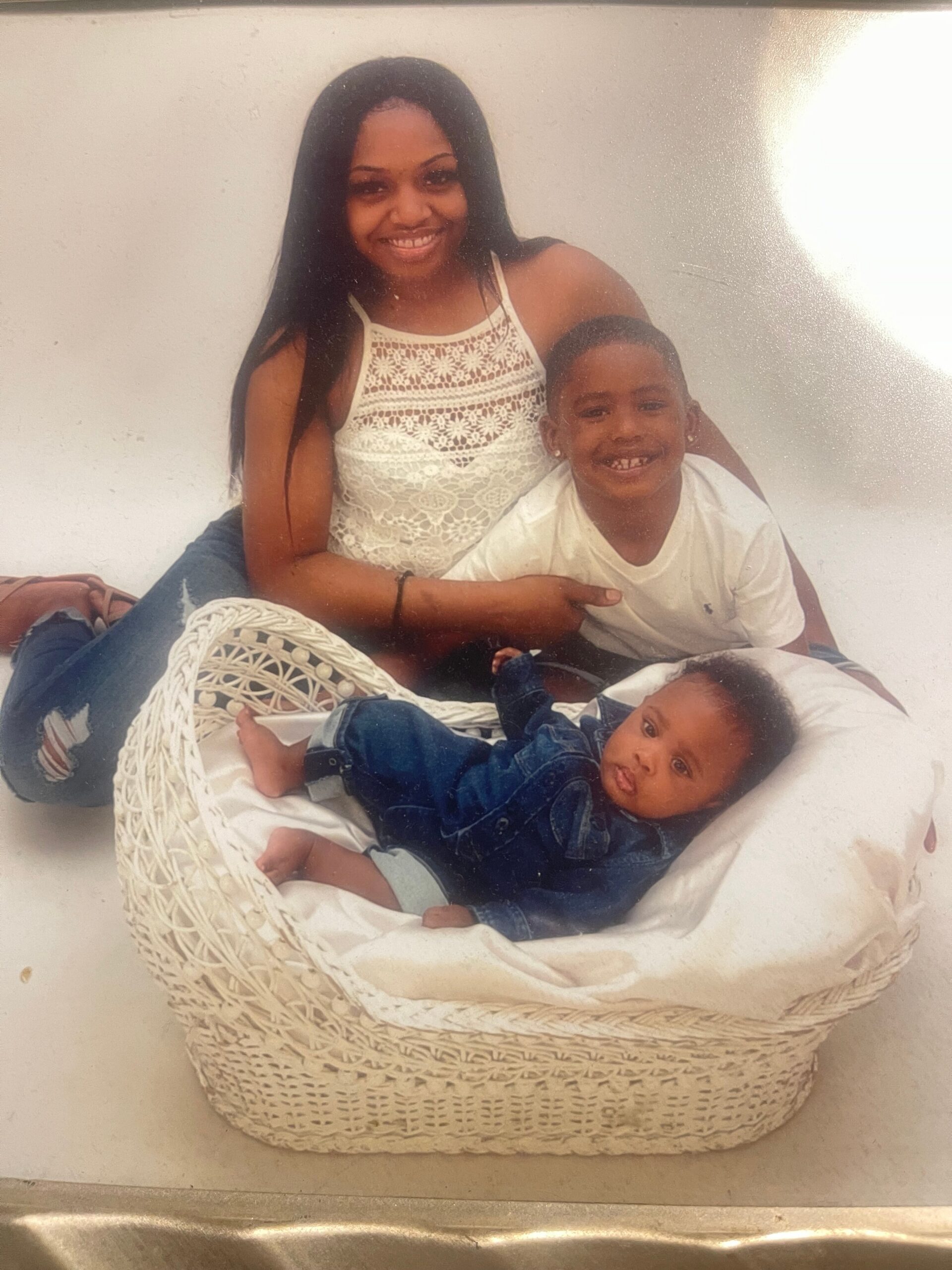 Smiling woman with long hair sits beside a toddler in a white lace top, all posing in a large woven basket with a baby in front.