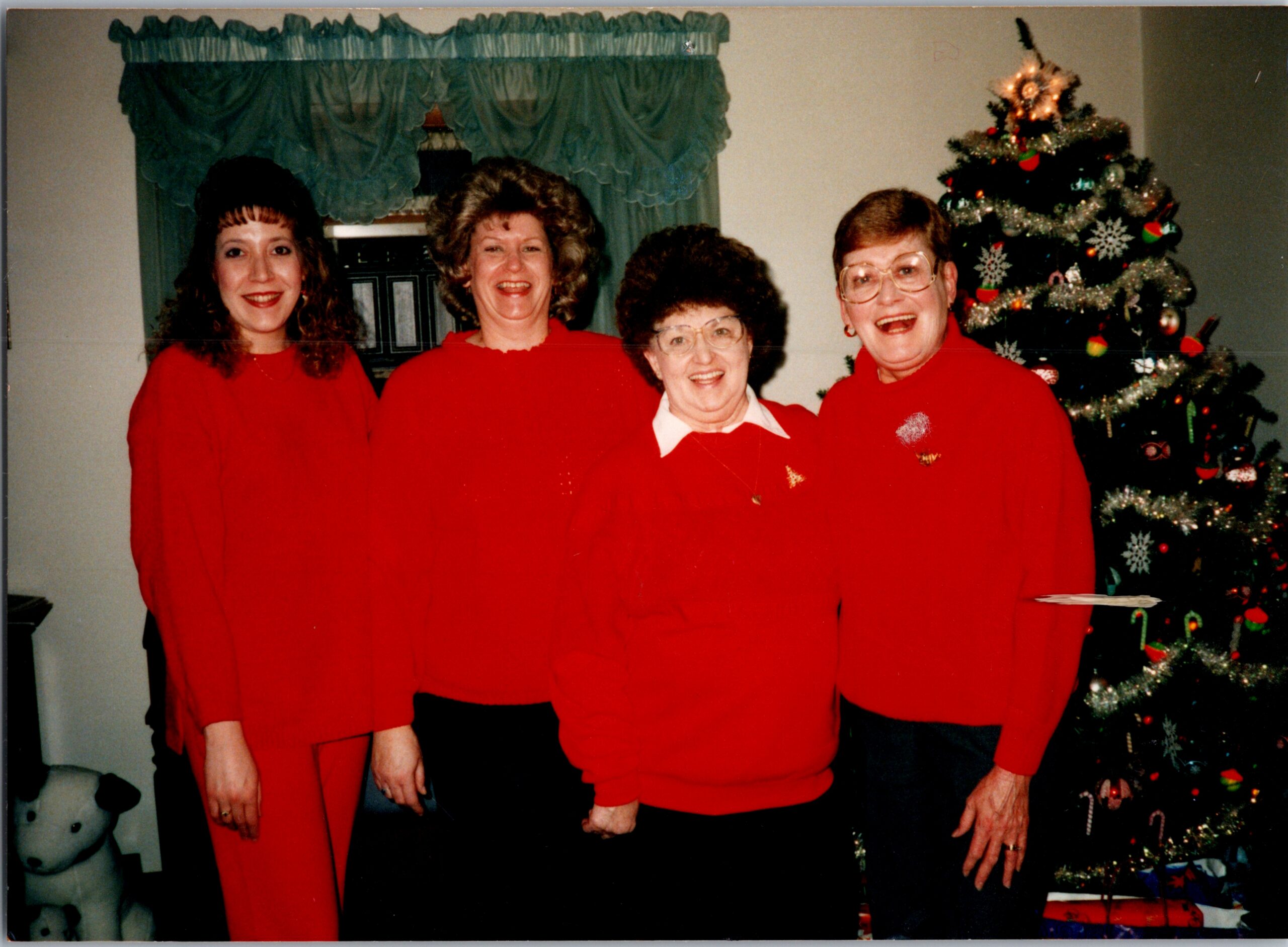 Four women in red sweaters posing together indoors for a Christmas photo with a decorated tree to the right.