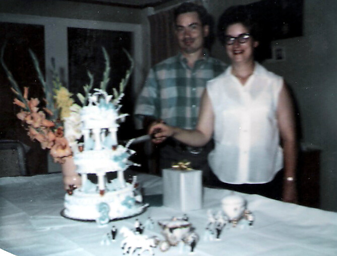 A man and smiling woman stand behind a white three‑tier cake on a table, cutting the cake at a home party.