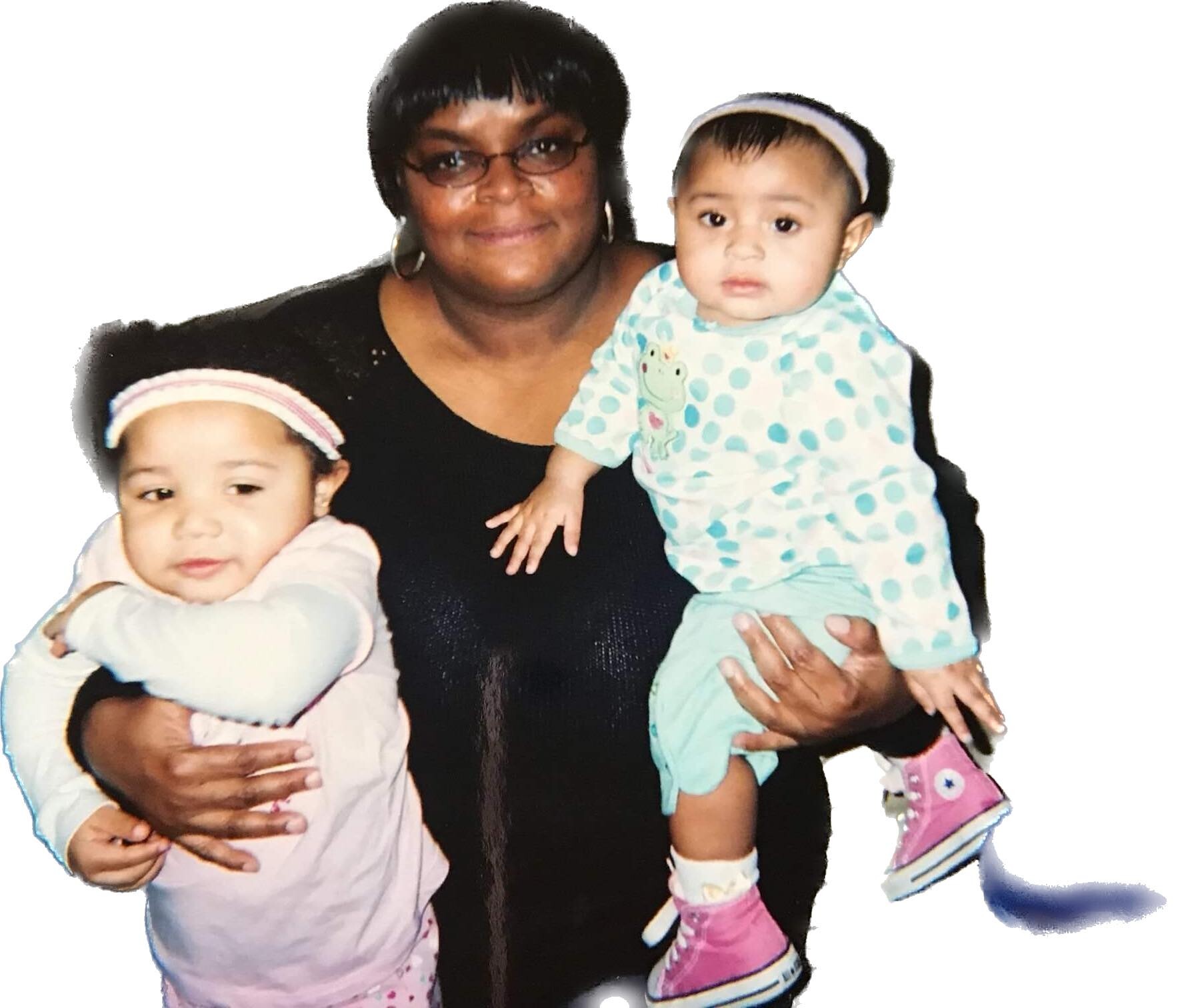 Woman in a black top holding two babies, one in pink on her left and one in turquoise polka-dot on her right, against a white background.