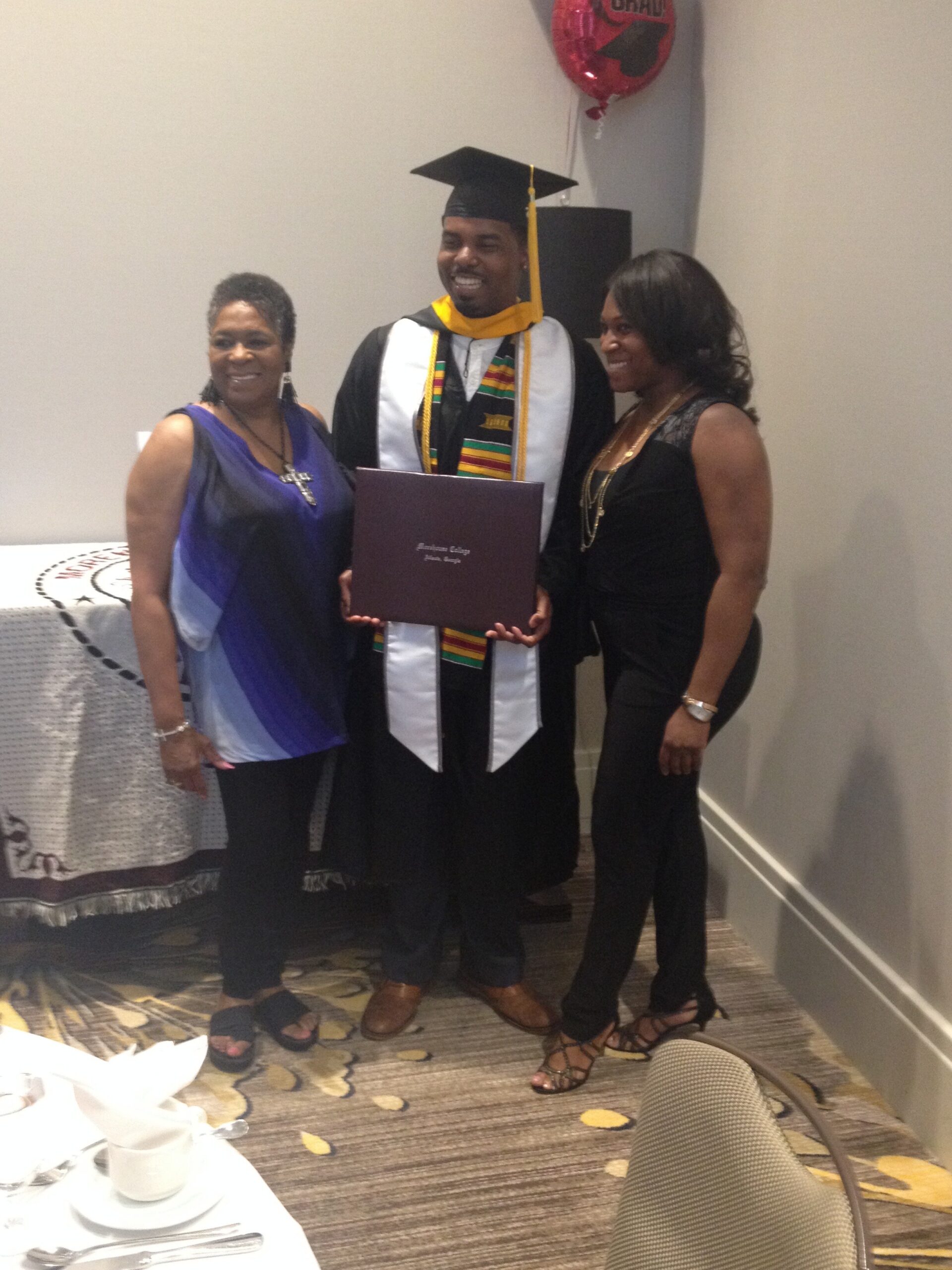 Graduate in cap and gown holding a purple diploma cover, flanked by two smiling women indoors.