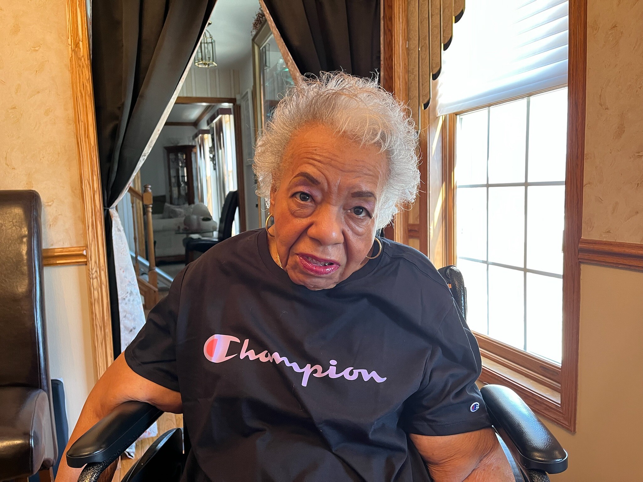 Older woman with short gray hair wearing a black Champion shirt, seated by a window in a wood-paneled room.