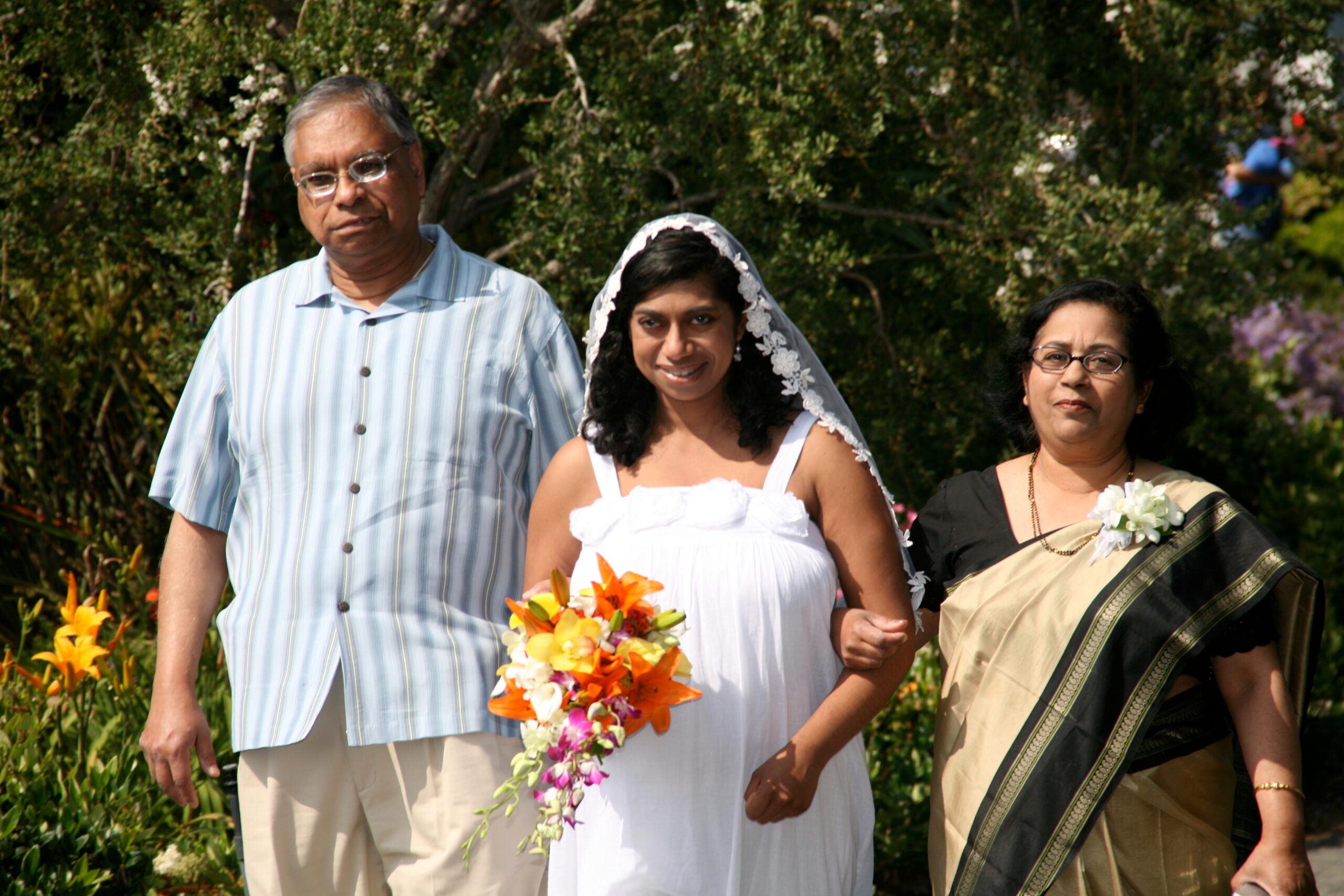Bride in white dress and veil walking outdoors with an older man in a striped shirt and an older woman in a sari, holding a colorful bouquet, greenery behind them.